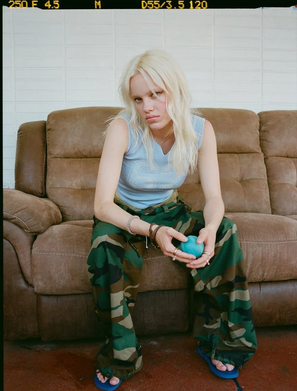 A young woman with long blonde hair, wearing a blue tank top and camo cargo pants, is sitting on a brown sofa holding a blue ball. She has multiple rings and bracelets and is sitting with her legs wide apart, wearing flip-flops.