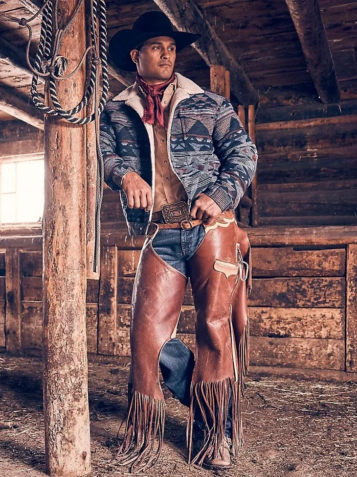 A man dressed as a cowboy in western attire, standing inside a rustic wooden barn.