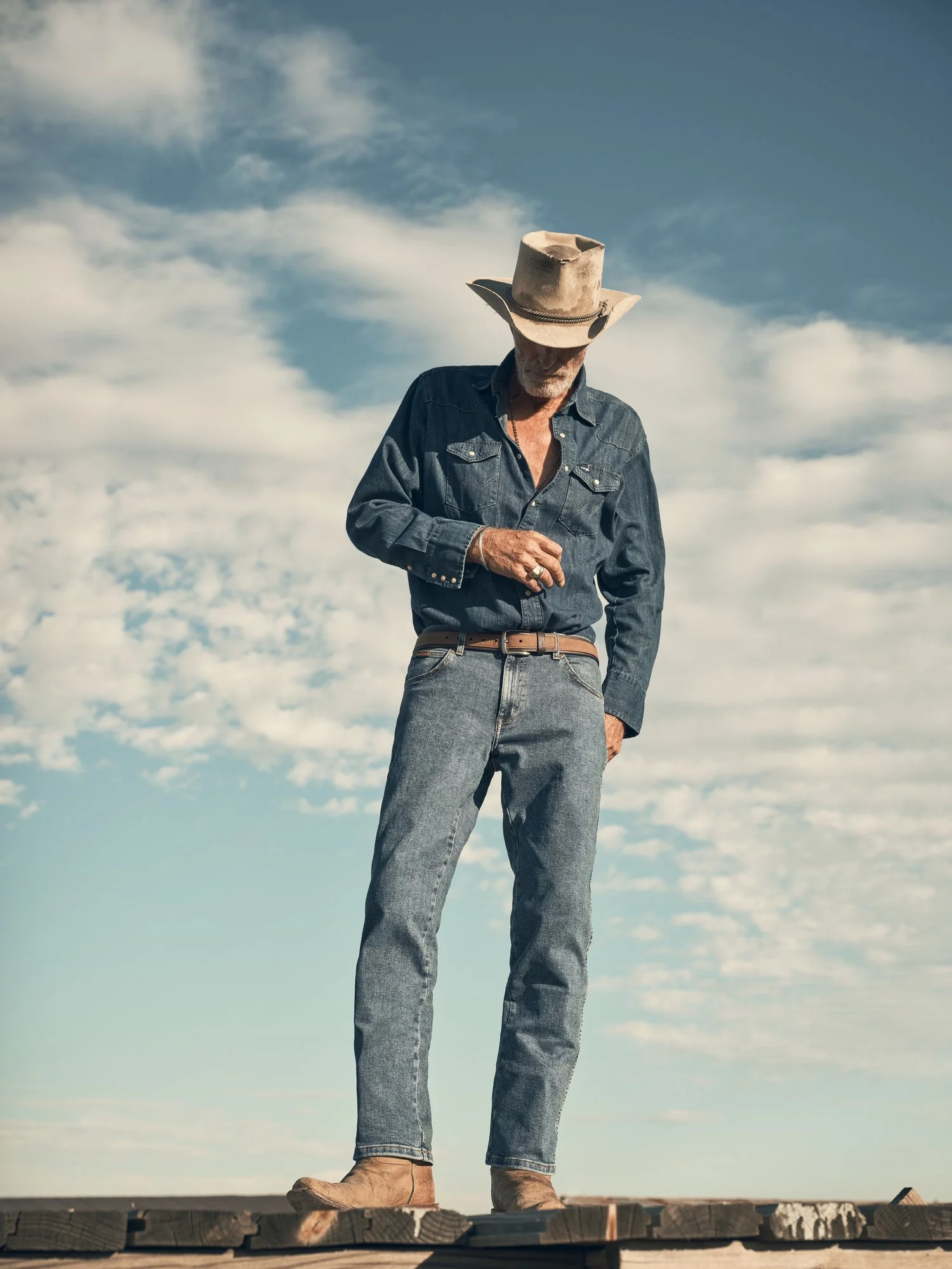 A man dressed in denim jeans and shirt, wearing a cowboy hat, standing outdoors on a wooden platform under a cloudy sky.