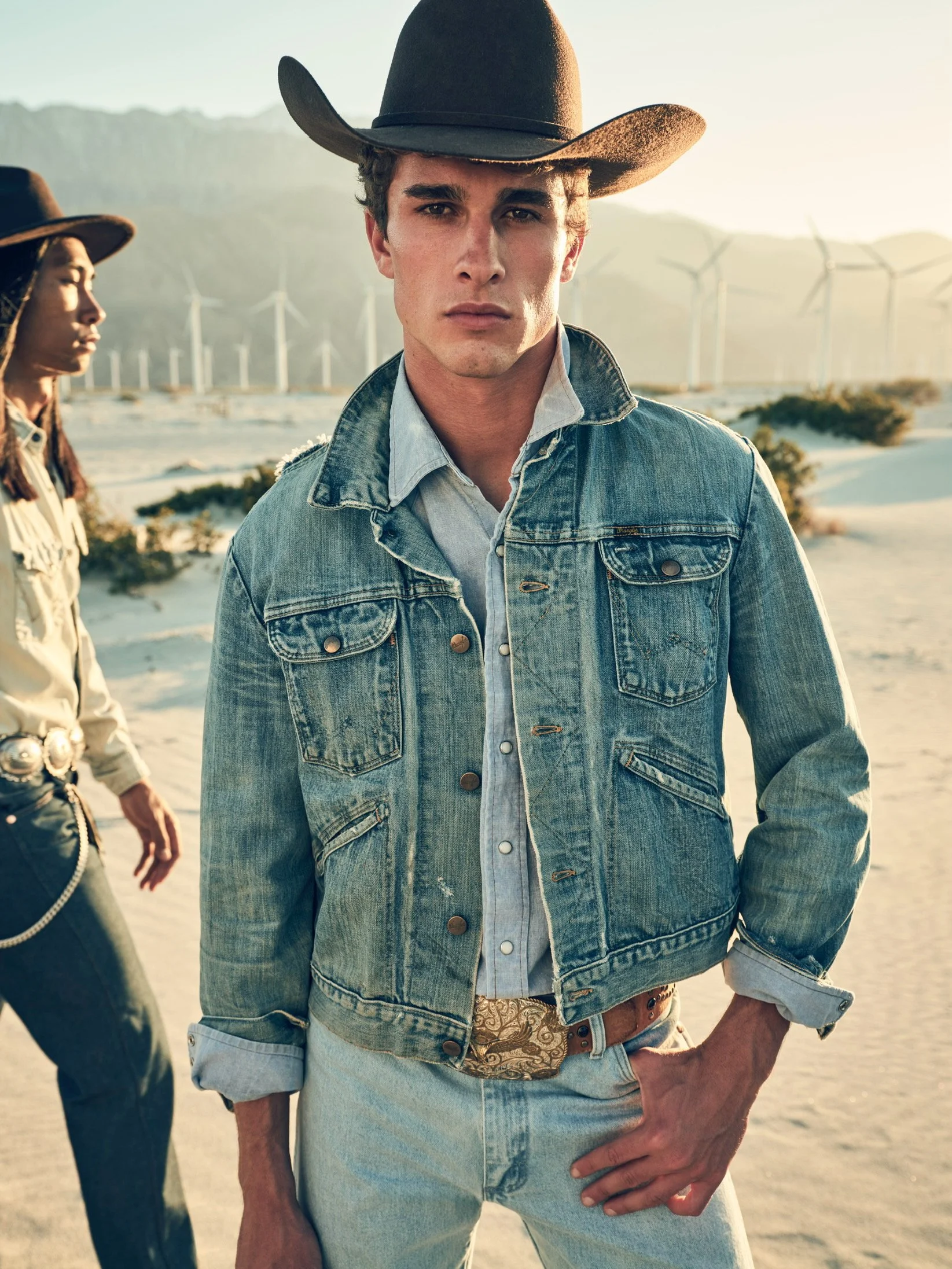 Young man wearing a cowboy hat and denim jacket standing in a desert landscape with wind turbines in the background.