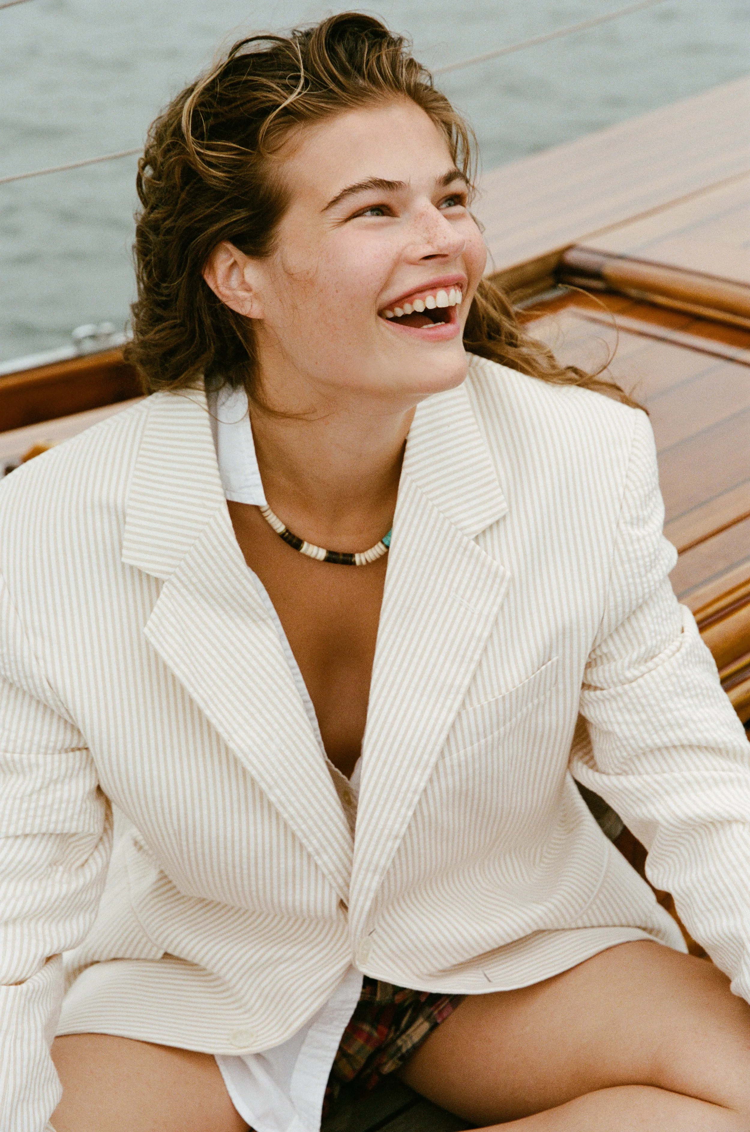 A young woman with curly hair, smiling and looking up, sitting on a wooden boat near water, wearing a striped blazer, a beaded necklace, and a plaid skirt.