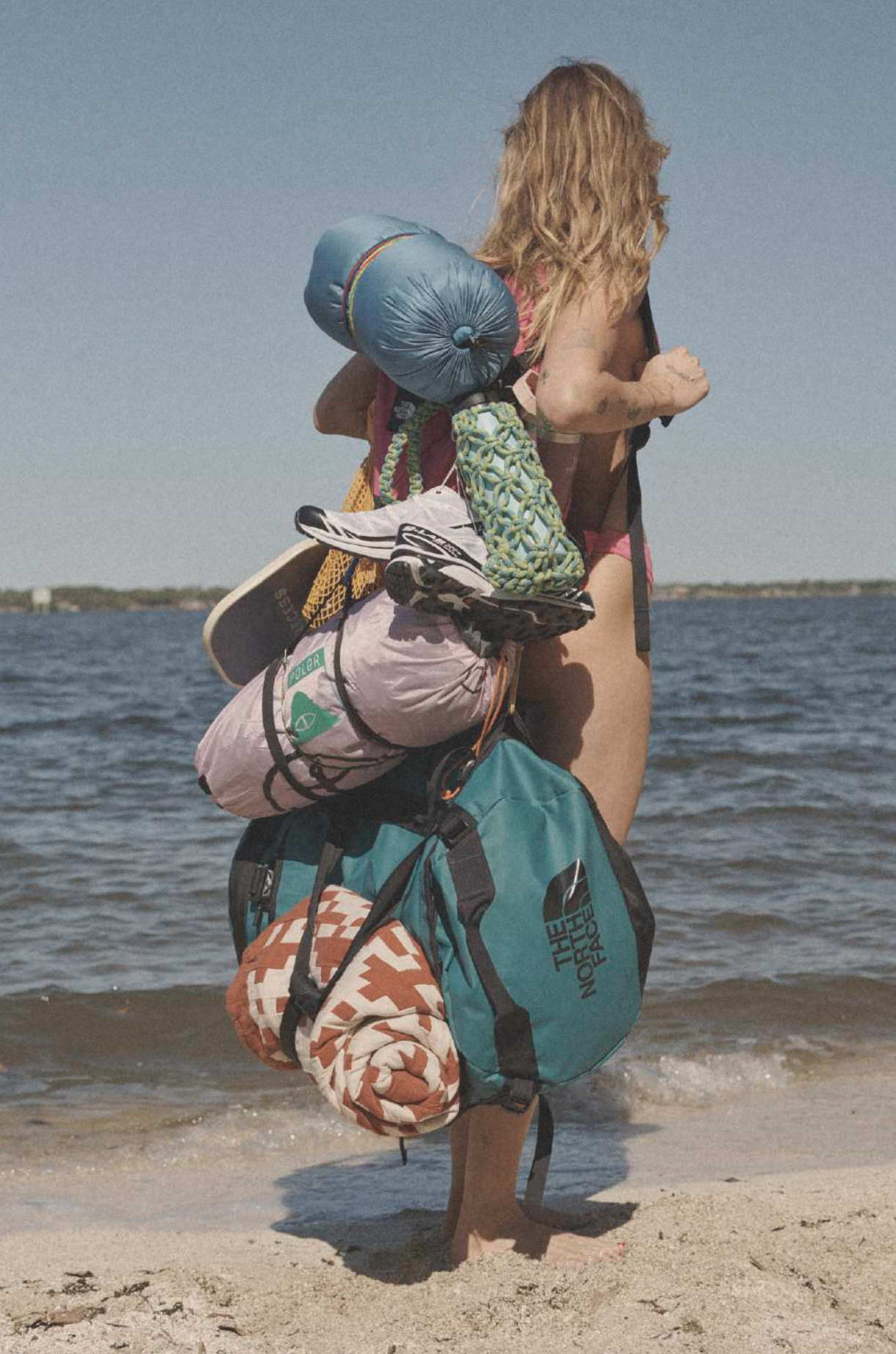 A woman with blonde hair wearing a bathing suit, carrying a large backpack filled with camping and outdoor gear, walking on a beach with water in the background.