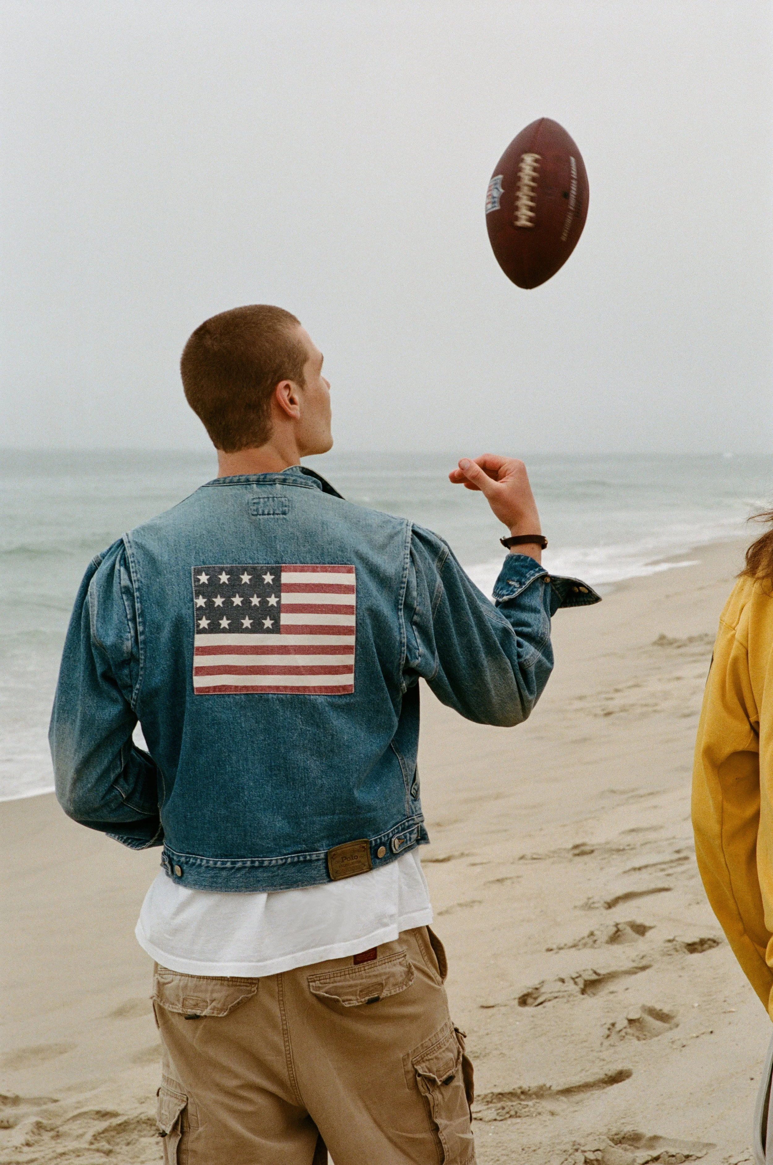 A young man in a denim jacket with an American flag patch on the back, standing on a sandy beach, tossing a football.