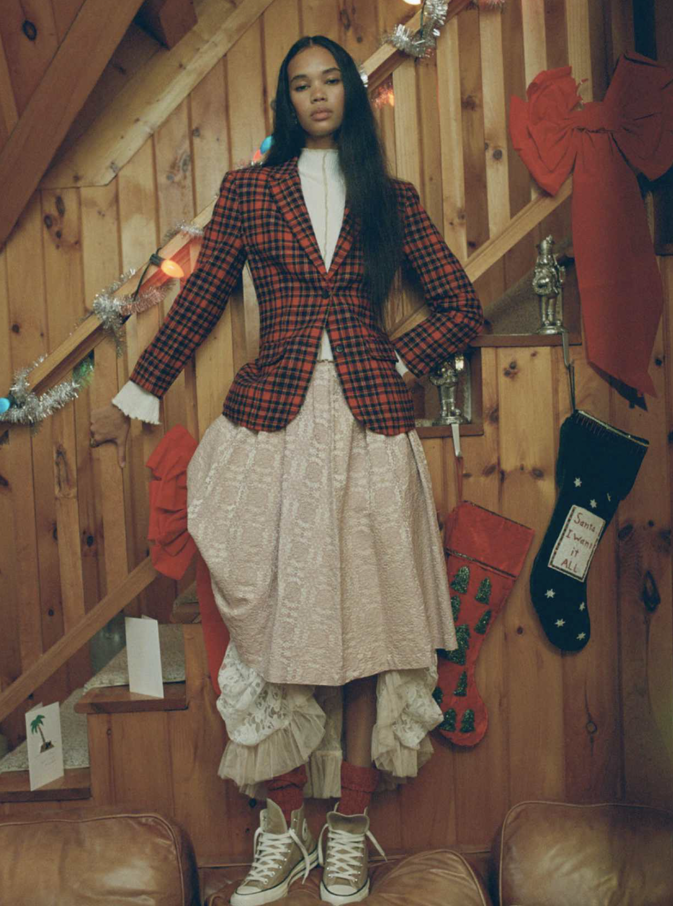 A woman standing on a couch in a decorated wooden staircase area, wearing a red plaid blazer, beige textured skirt with ruffled layers, beige sneakers, and red socks, with Christmas decorations and stockings in the background.