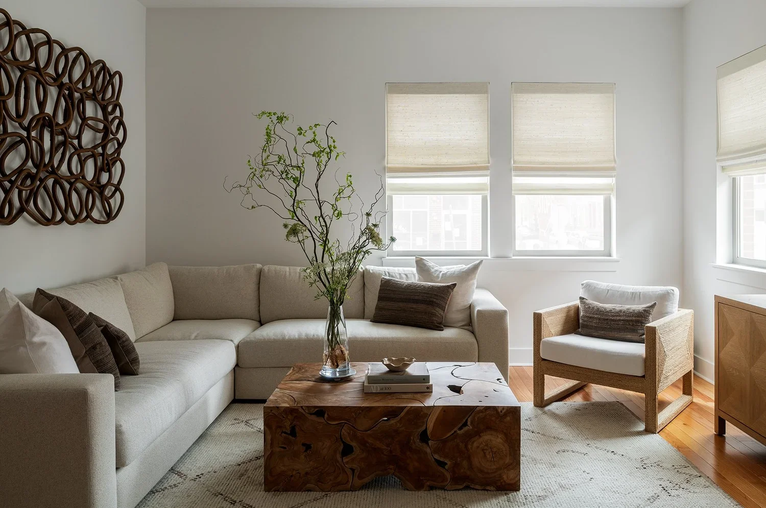 A warm, contemporary living room designed with natural textures, organic forms, and a calming neutral palette. A large L-shaped sectional sofa upholstered in soft ivory fabric anchors the space, layered with textured accent pillows in shades of taupe
