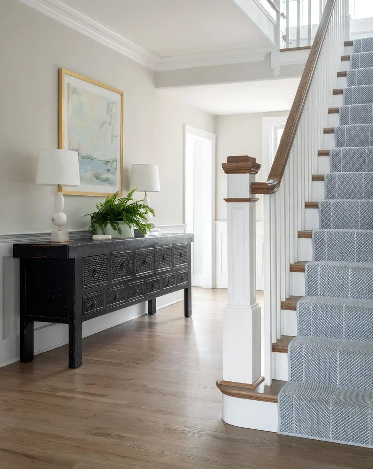 Luxury transitional foyer design by Kate Kelly Interiors featuring custom millwork, white wainscoting, a black console table, blue stair runner, and white oak flooring in a timeless Pennsylvania home.