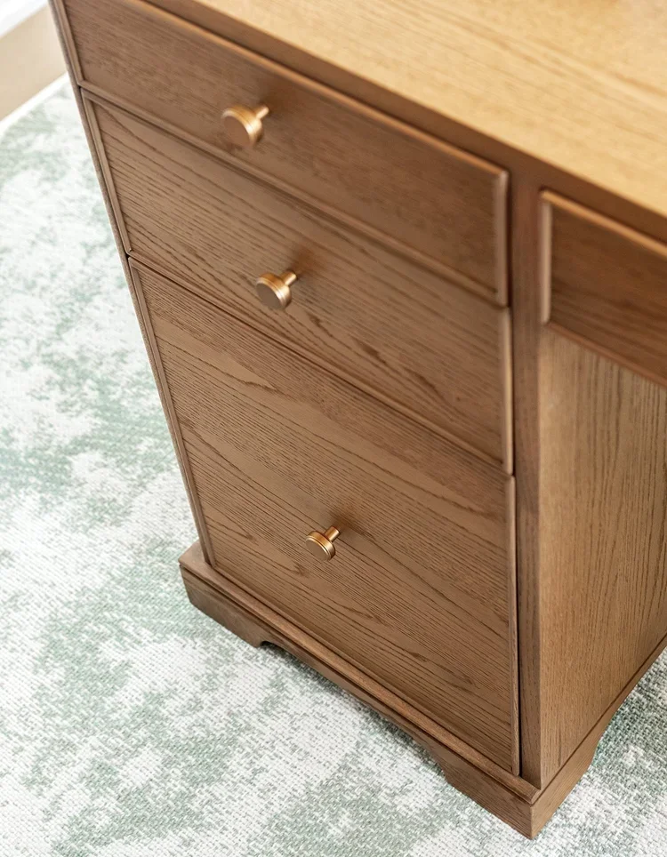 A close-up detail of a custom wood desk pedestal, showcasing the rich grain and warm tones of stained white oak cabinetry. Three seamlessly integrated drawers are fitted with brushed brass round knobs, creating a refined balance between modern simpli