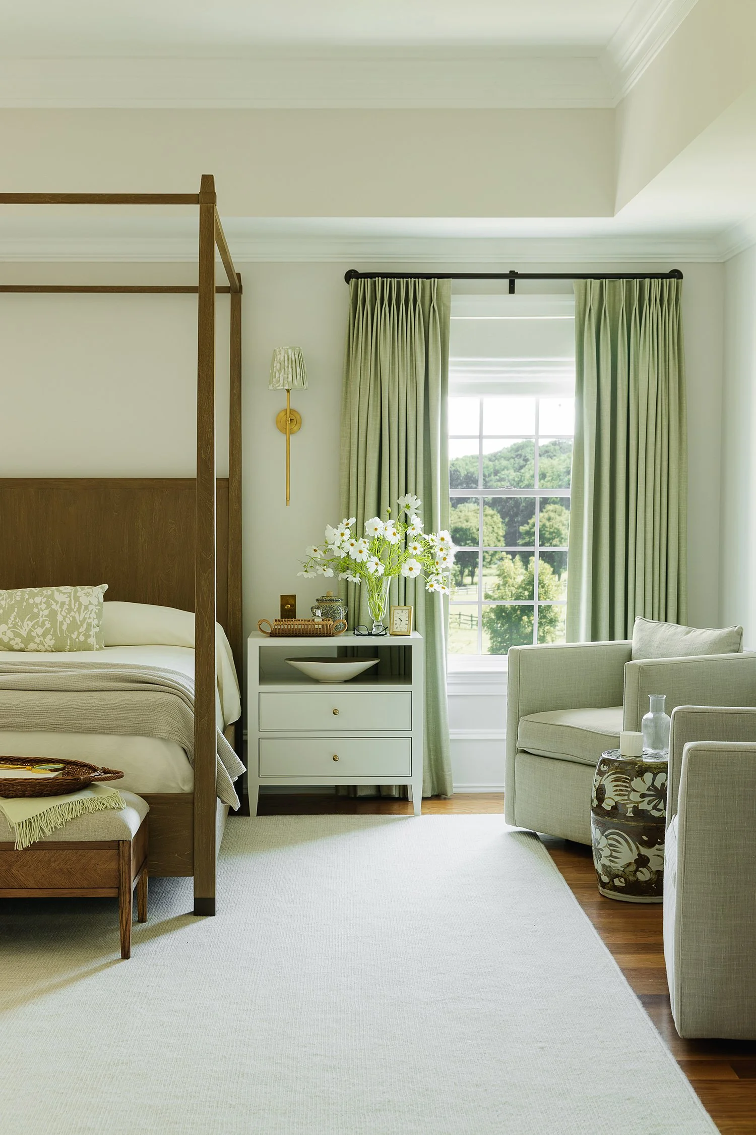 Bright luxury primary suite featuring a walnut canopy bed, custom green drapery, elegant white nightstand, upholstered lounge chairs, textured area rug, and abundant natural light overlooking scenic landscape views.
