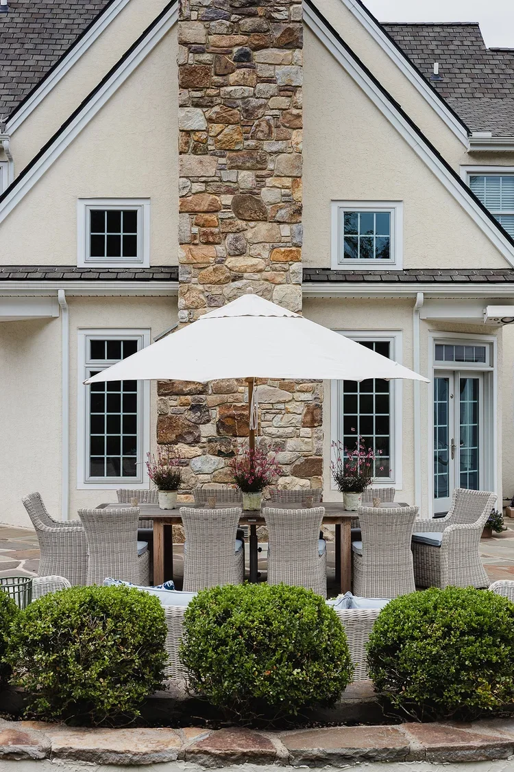 An elegant outdoor dining terrace designed for both entertaining and everyday relaxation. Centered against the home's cream-colored stucco exterior, a substantial natural stone chimney creates a striking architectural focal point. A large dining tabl