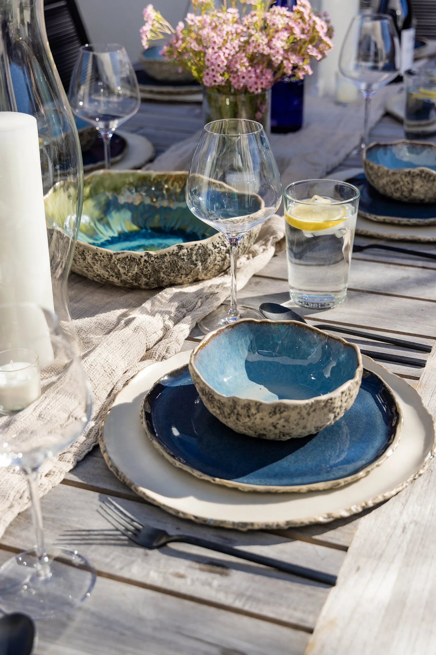 Elegant outdoor dining table styled with handcrafted blue ceramic dinnerware, wine glasses, linen textiles, candles, and fresh flowers. Coastal-inspired tablescape featuring layered textures, artisan pottery, and sophisticated alfresco entertaining.