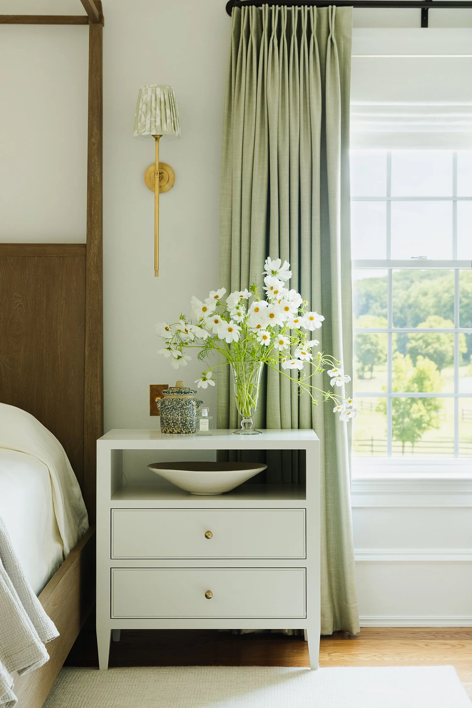 Luxury primary bedroom featuring a custom white nightstand styled with fresh white cosmos flowers, decorative ceramics, brass wall sconce, tailored green linen drapery, and a walnut canopy bed, creating a timeless and elegant retreat by Kate Kelly In