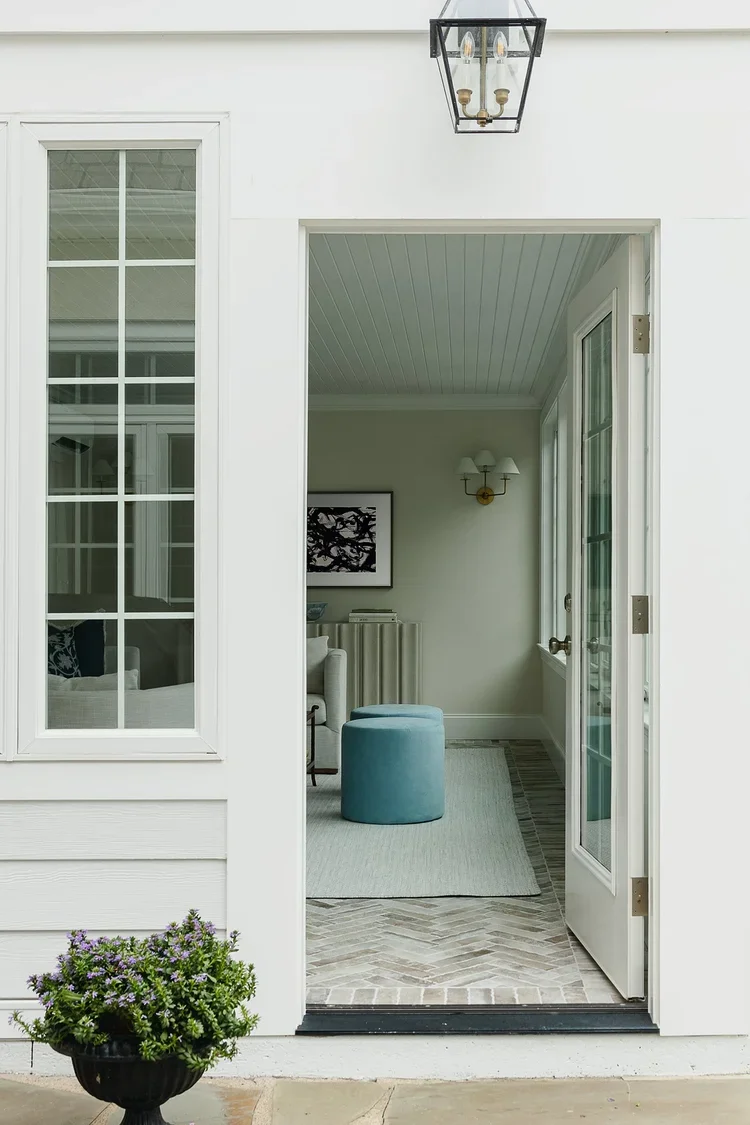 The transition between an outdoor patio and an inviting covered porch or sunroom, emphasizing the home's seamless indoor-outdoor connection. The exterior features crisp white siding, classic divided-light windows, and a black metal lantern mounted ab