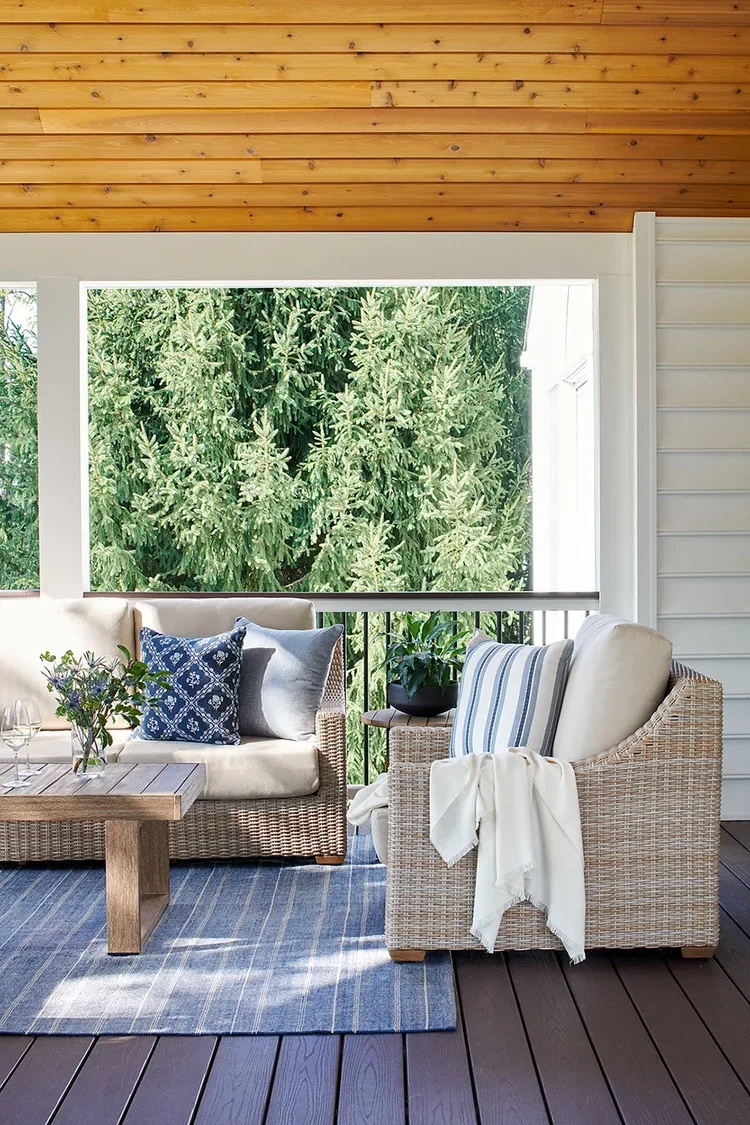 A sophisticated covered porch designed for comfort, relaxation, and year-round outdoor living. Woven wicker lounge seating with plush ivory cushions creates an inviting conversation area, accented by layered blue and white patterned pillows that intr