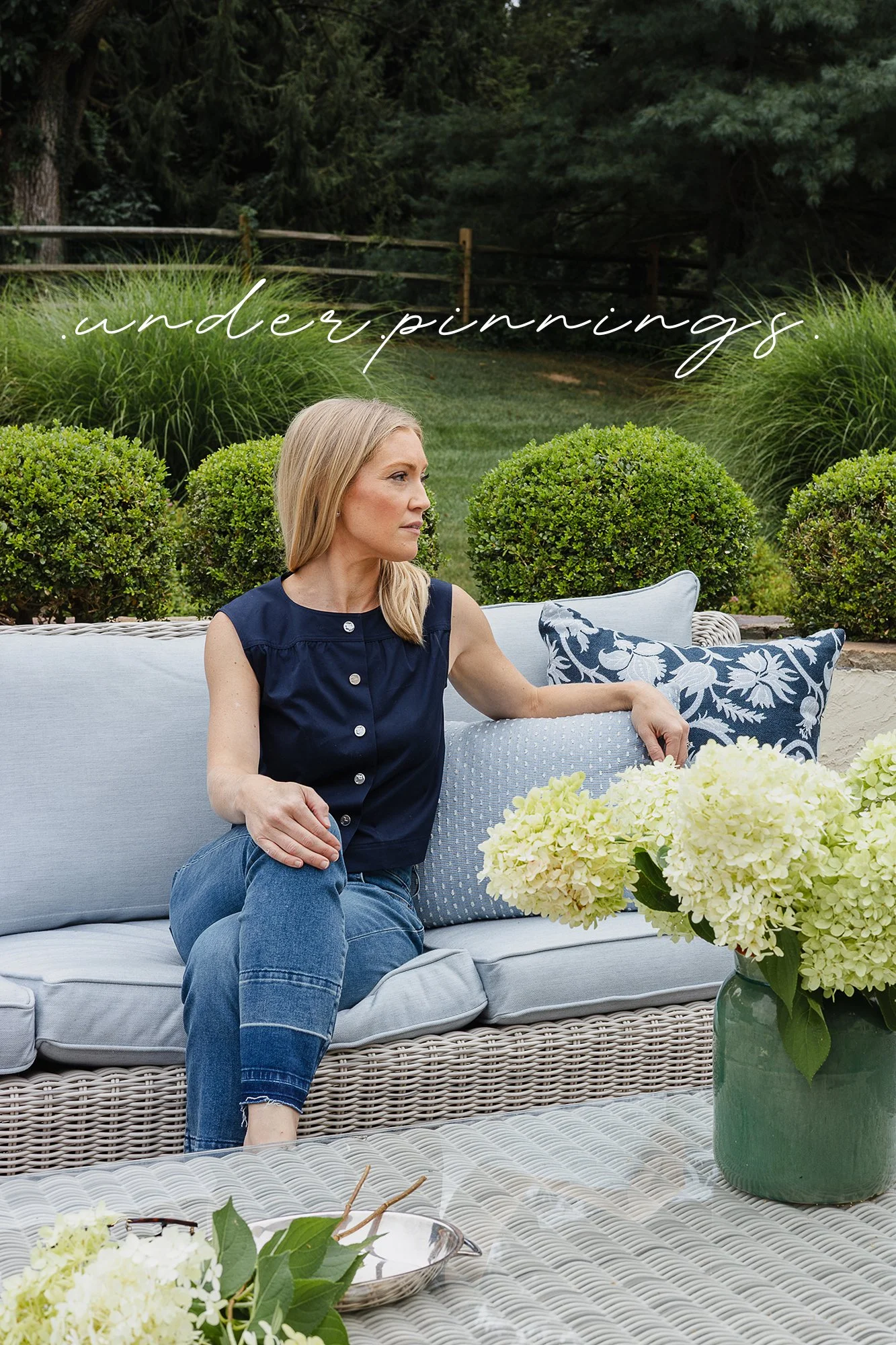 Interior designer seated on a woven outdoor sofa surrounded by hydrangeas and manicured greenery. Elegant outdoor living space with layered textiles, garden landscaping, and timeless coastal-inspired design for luxury residential living.