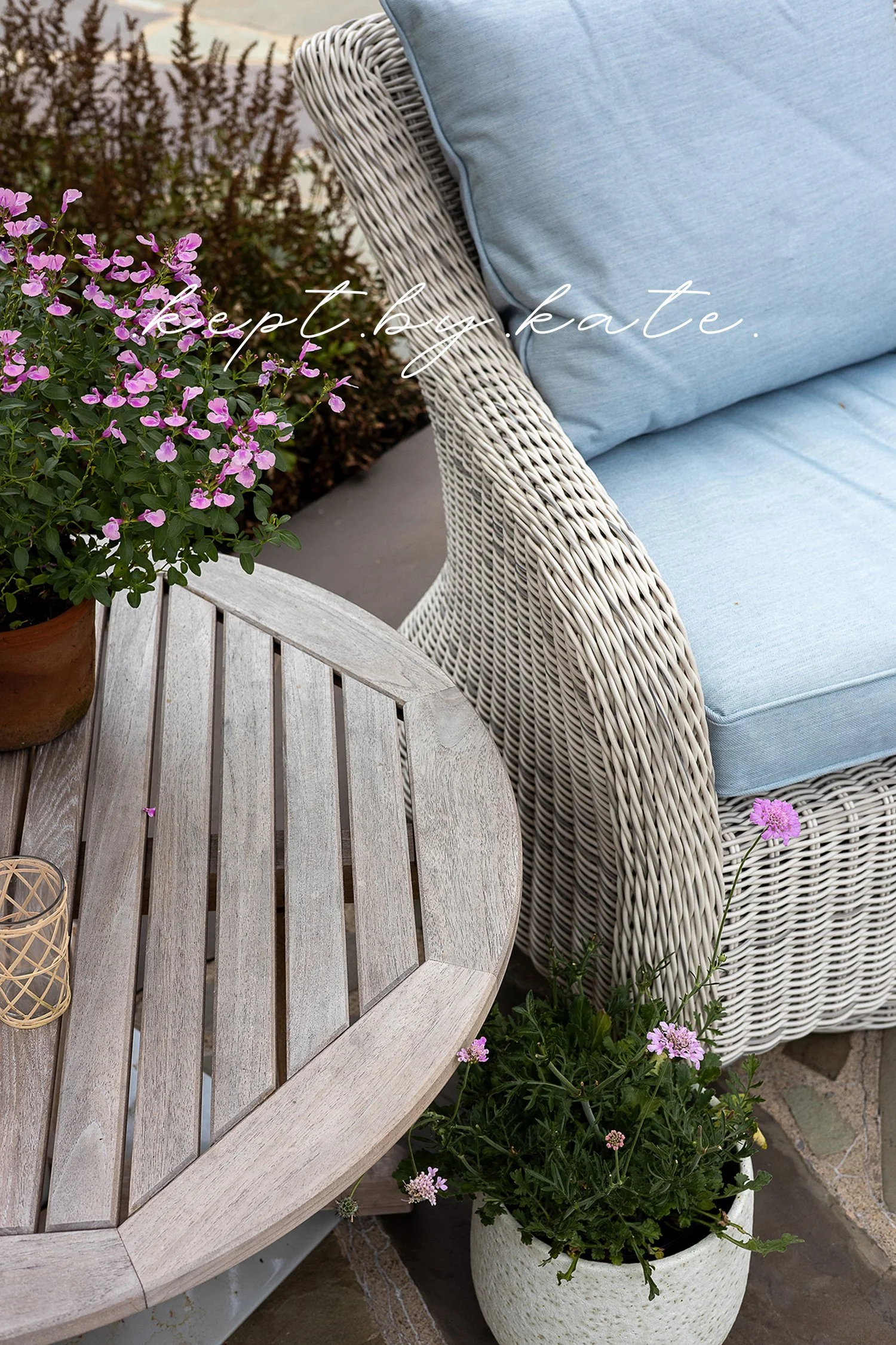 Close-up of a luxury outdoor seating area featuring a woven wicker lounge chair, pale blue cushions, weathered wood table, and flowering potted plants. Elegant garden patio design with layered textures, soft colors, and timeless outdoor living style.