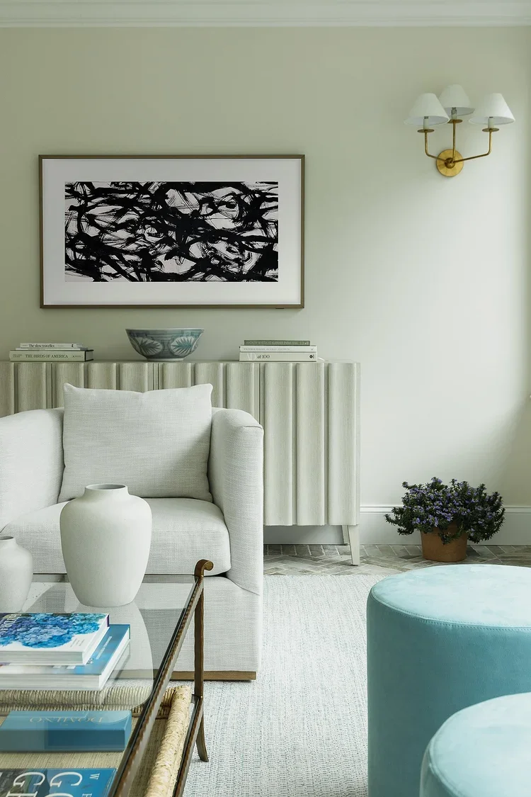 A sophisticated seating vignette within a bright transitional sunroom, showcasing a carefully curated blend of comfort, texture, and refined design. A plush white upholstered lounge chair anchors the foreground, paired with a glass-top coffee table s