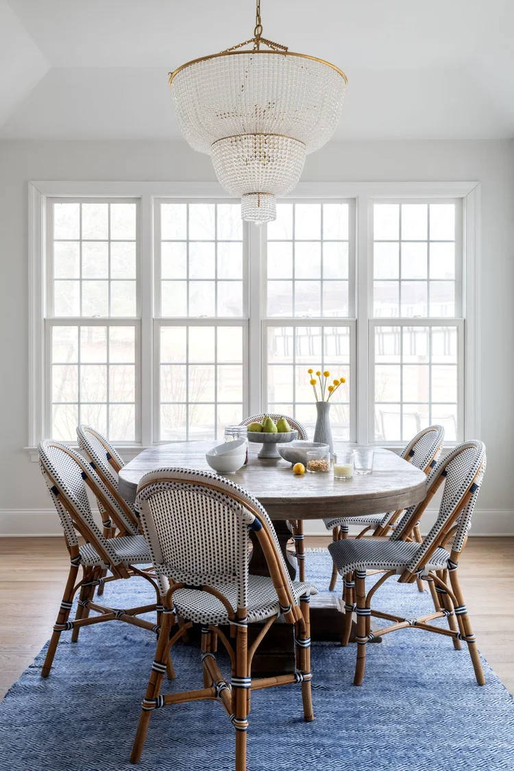 Bright transitional dining room featuring a long, rounded wood dining table, woven bistro chairs, a crystal chandelier, blue area rug, and large windows flooding the space with natural light.