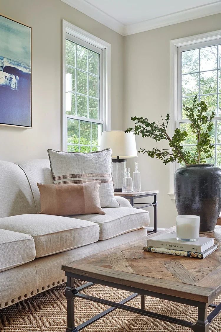 A sophisticated transitional living room vignette centered around layered textures, natural materials, and abundant daylight. A cream upholstered sofa with nailhead trim is styled with oversized linen pillows featuring subtle stripe detailing and a s