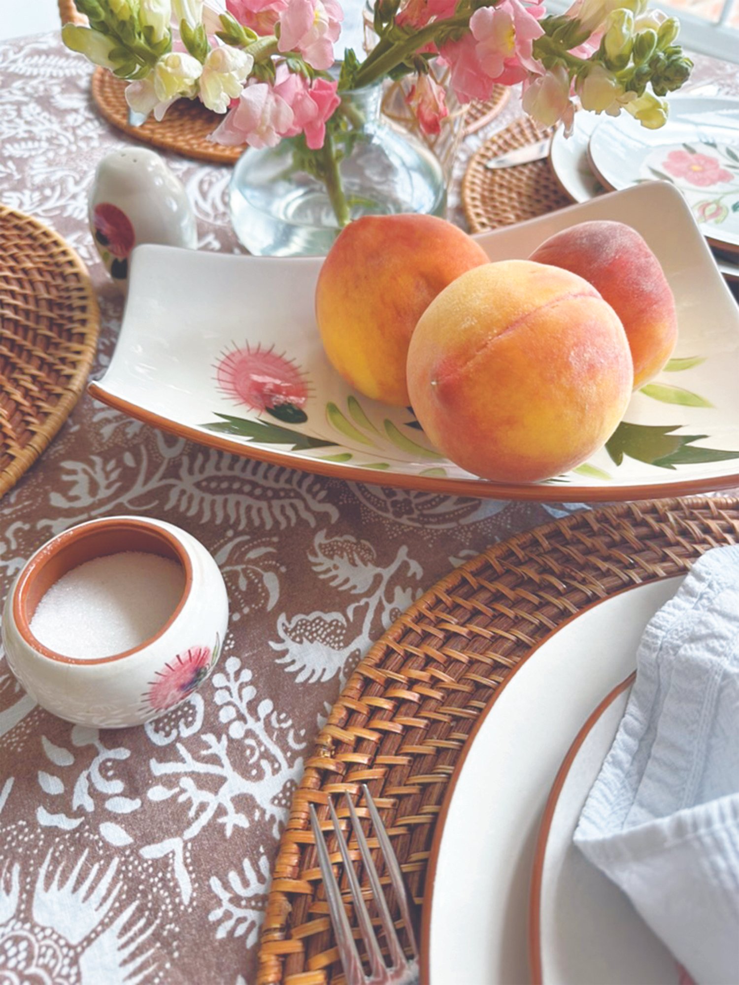 Summer tablescape with fresh peaches, pink snapdragons, rattan placemats, hand-painted ceramic serveware, and a botanical tablecloth.