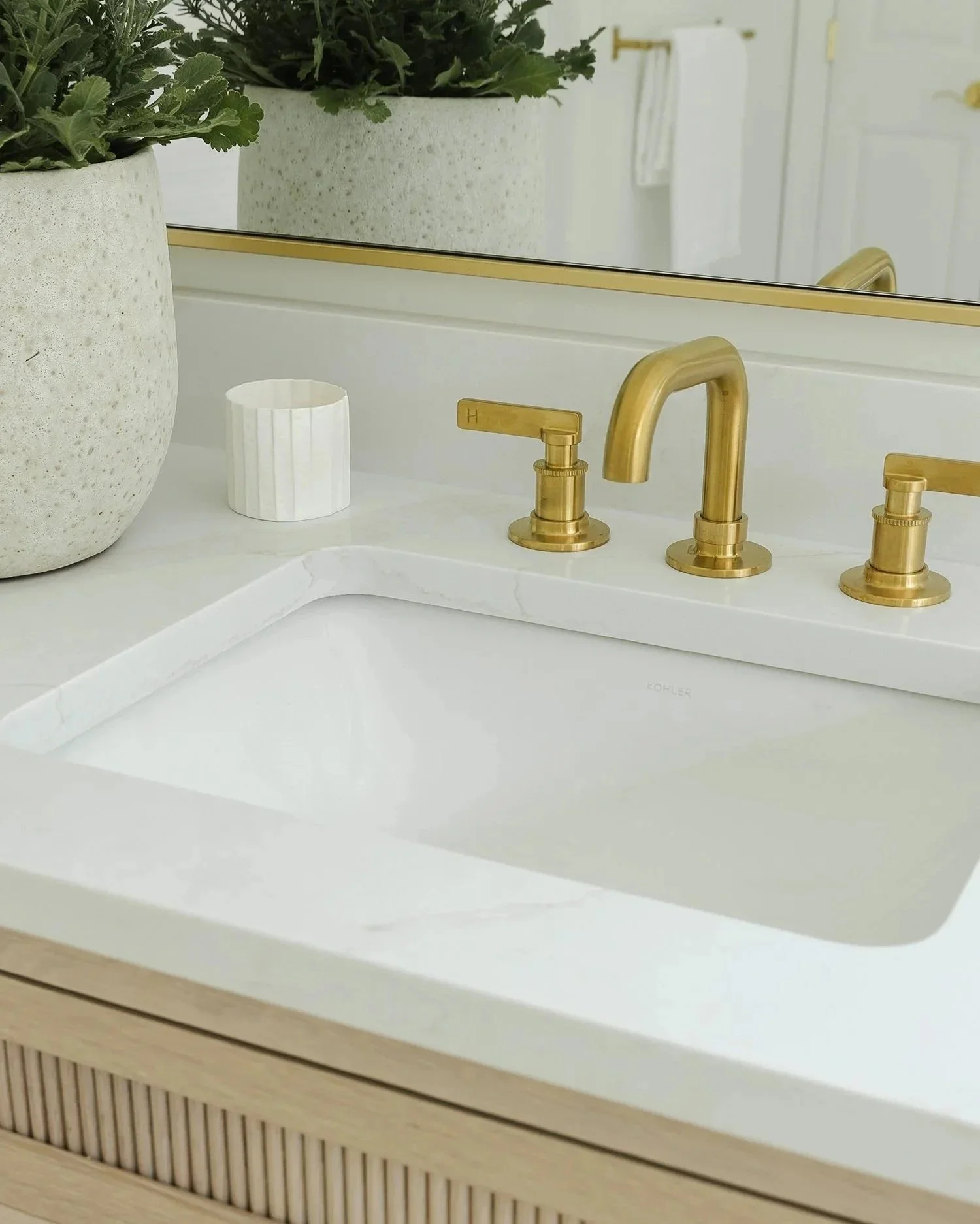 Modern bathroom vanity with a white quartz integrated sink, brushed brass faucet, brass-framed mirror, and light oak fluted cabinetry. Green plants add warmth to the bright, minimalist space, creating a luxurious spa-inspired modern coastal design.