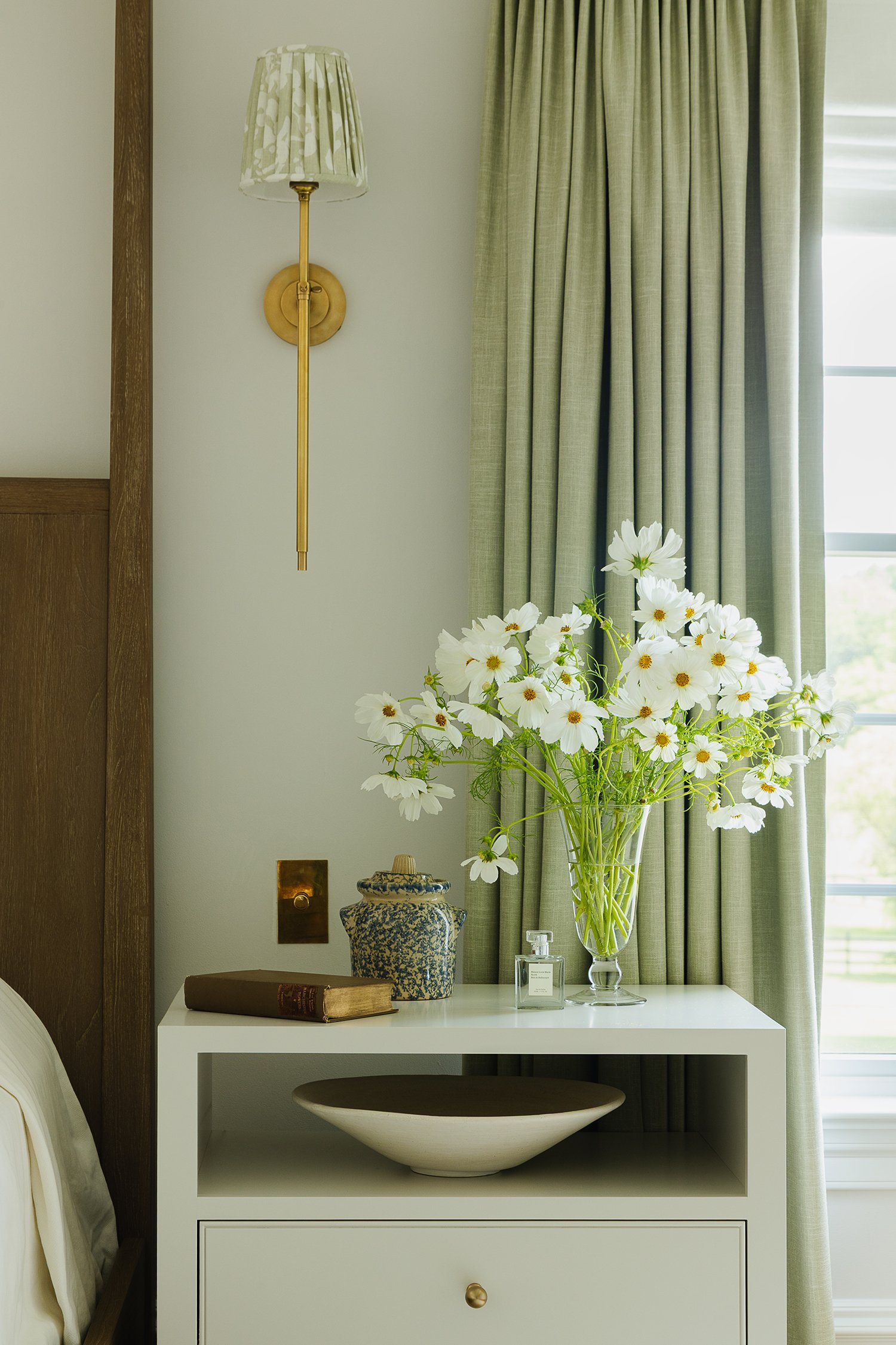 Elegant white bedside table styled with fresh white flowers, decorative ceramics, vintage books, brass hardware, and custom linen drapery beside a walnut canopy bed in a refined primary suite.