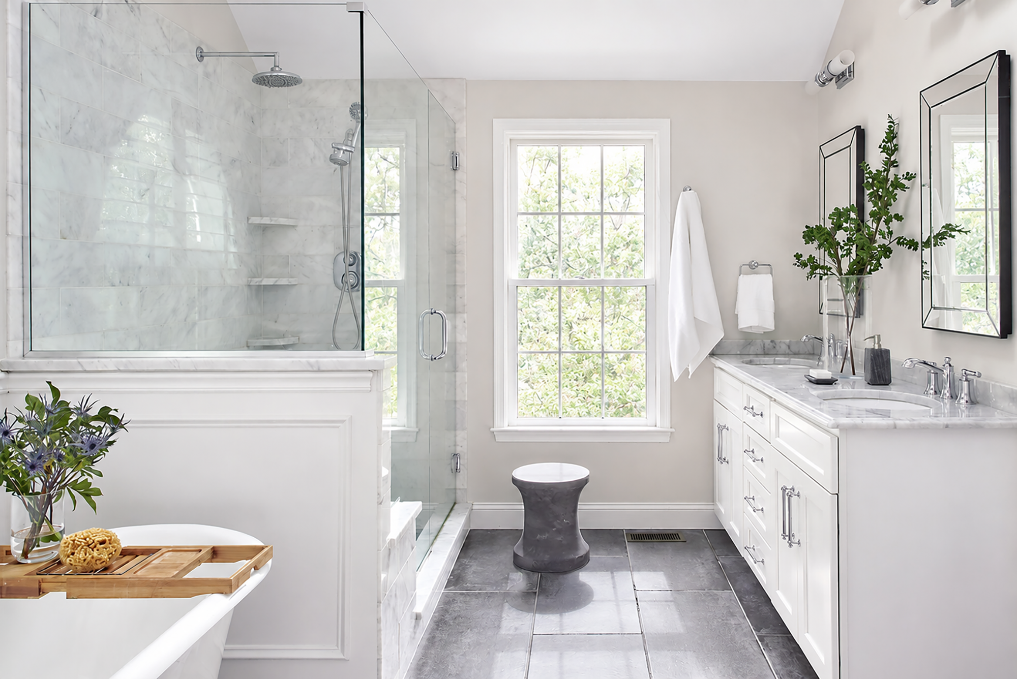 Luxury bathroom featuring a glass-enclosed marble shower, double vanity with marble countertop, freestanding tub, and large windows. Bright transitional design with chrome fixtures, gray tile flooring, and elegant spa-inspired finishes.