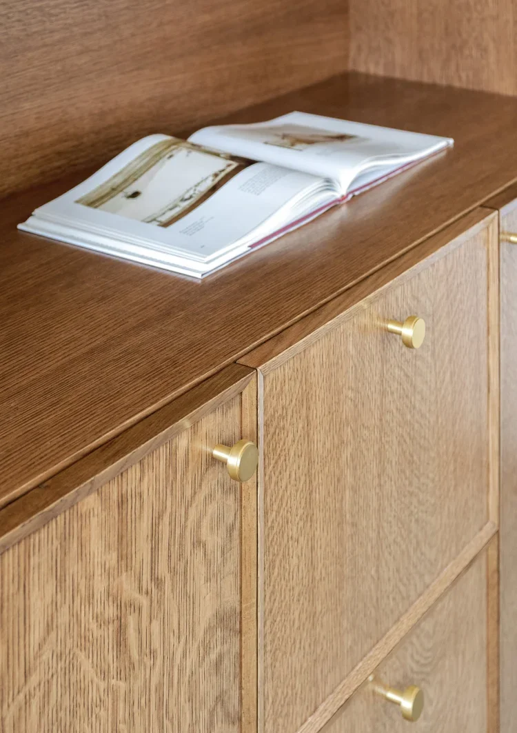 A close-up detail of custom white oak cabinetry, highlighting the natural wood grain and fine craftsmanship of the millwork. The cabinet doors feature simple recessed paneling and are accented with warm brushed brass round knobs that add a subtle tou
