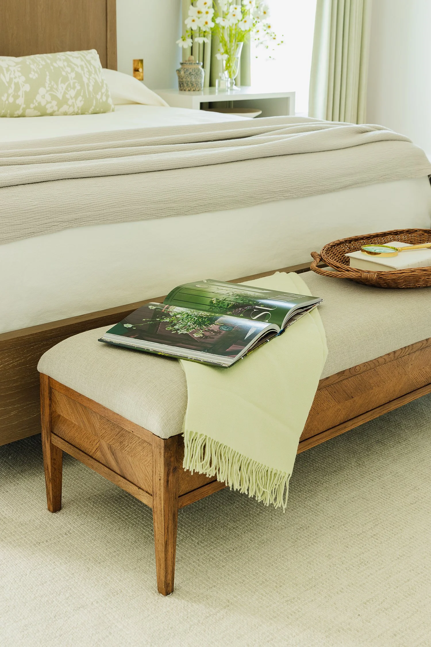 Upholstered wood bench at the foot of a luxury bed styled with a soft throw, design book, and woven tray in a serene primary bedroom featuring layered neutral bedding and custom furnishings.