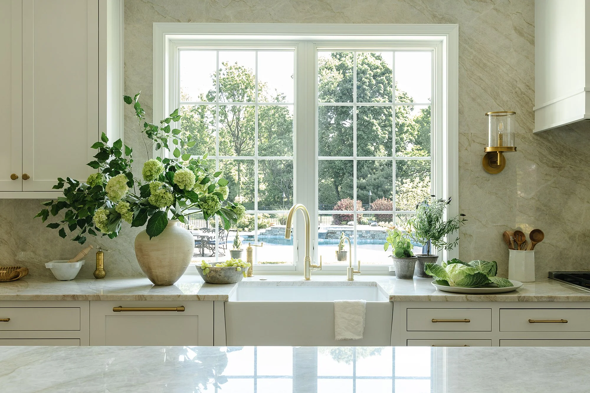 Luxury kitchen with a farmhouse sink beneath a large window, brass faucet, stone slab backsplash, white cabinetry, and fresh greenery. Natural light highlights the timeless transitional design, custom millwork, and elegant finishes.