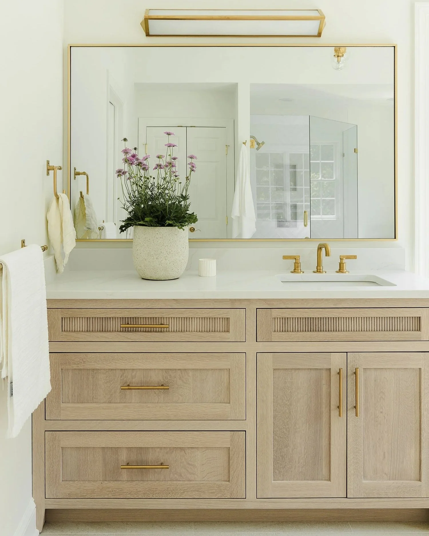 Modern bathroom vanity with light oak cabinetry, brass hardware, white quartz countertop, and integrated sink. A brass-framed mirror, matching faucet, and potted flowers create a bright, elegant, spa-inspired bathroom with modern coastal style.