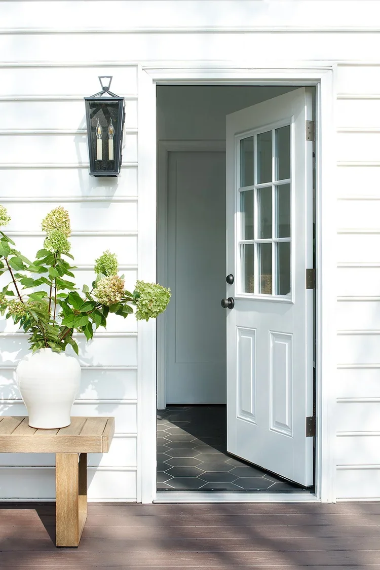 A welcoming exterior entry framed by crisp white horizontal siding and classic architectural details. A partially open white Dutch-style door with divided glass panes creates an inviting transition between the outdoor living space and the home's inte