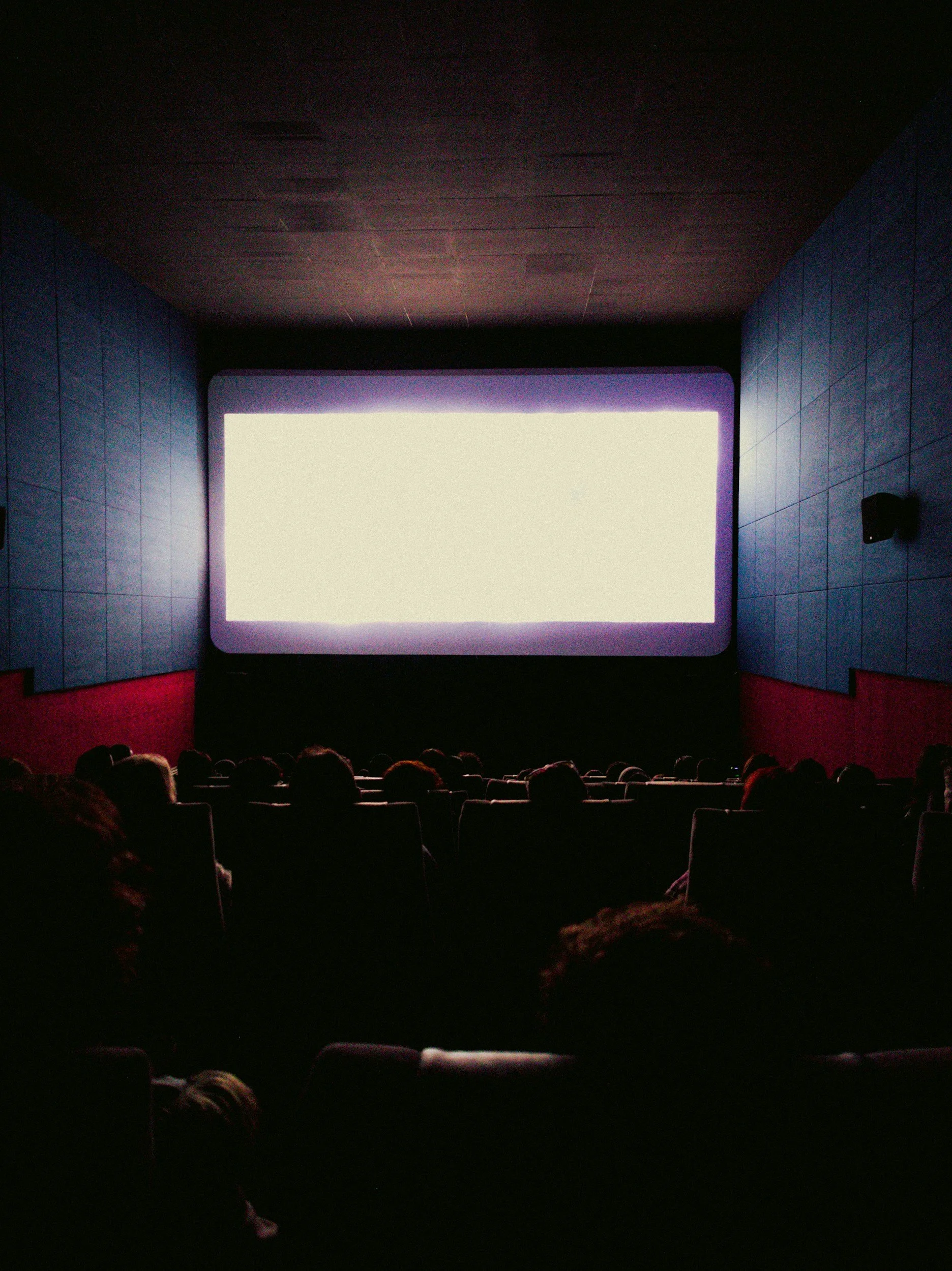 A dark movie theater with silhouettes of people watching a bright screen, showing the backs of their heads and setting with blue walls and red curtains.