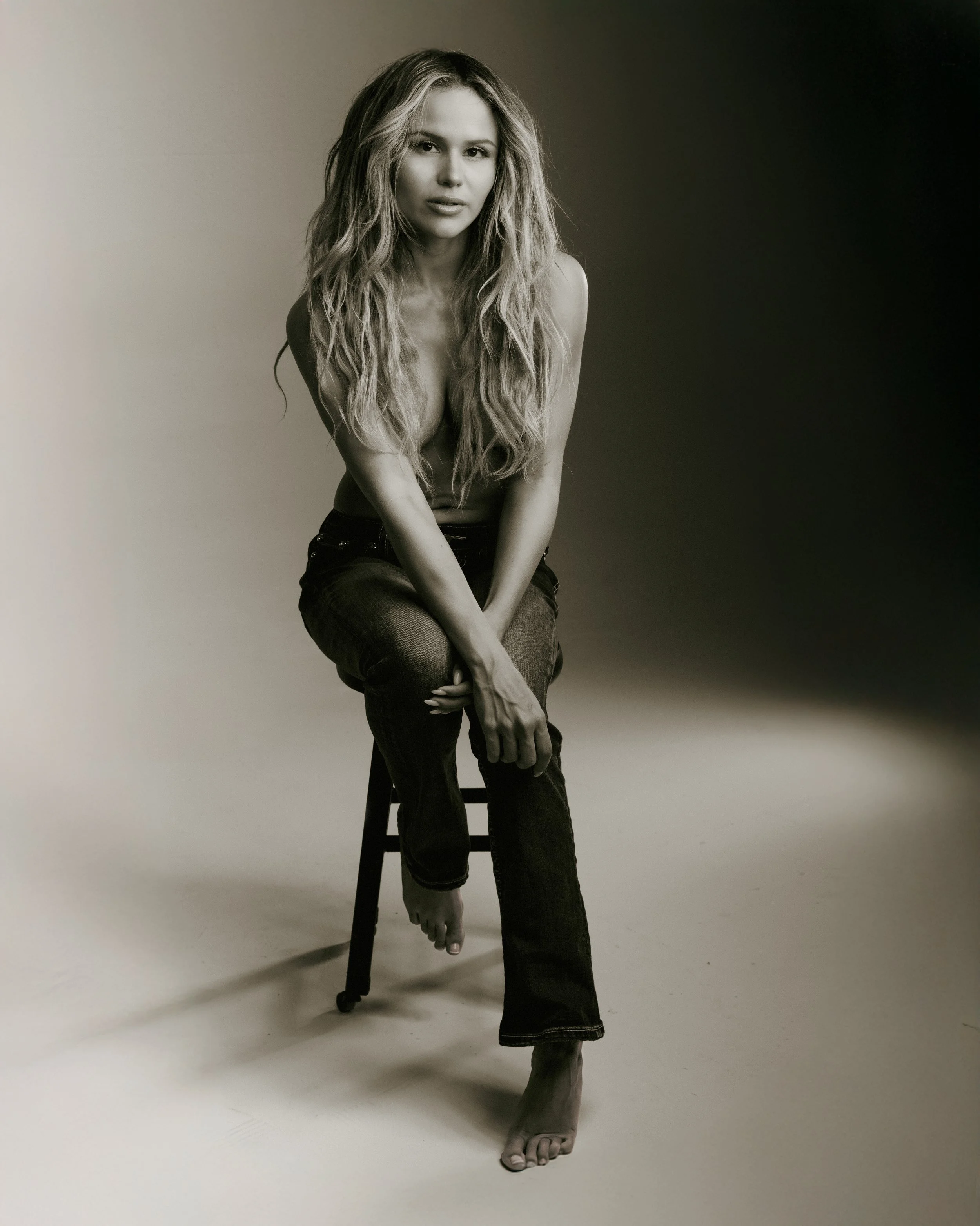 A beautiful black and white photo by expert LA fashion and portrait photographer Joe Fortunato of a model with long wavy hair sitting on a stool, wearing jeans, indoors with a plain background.