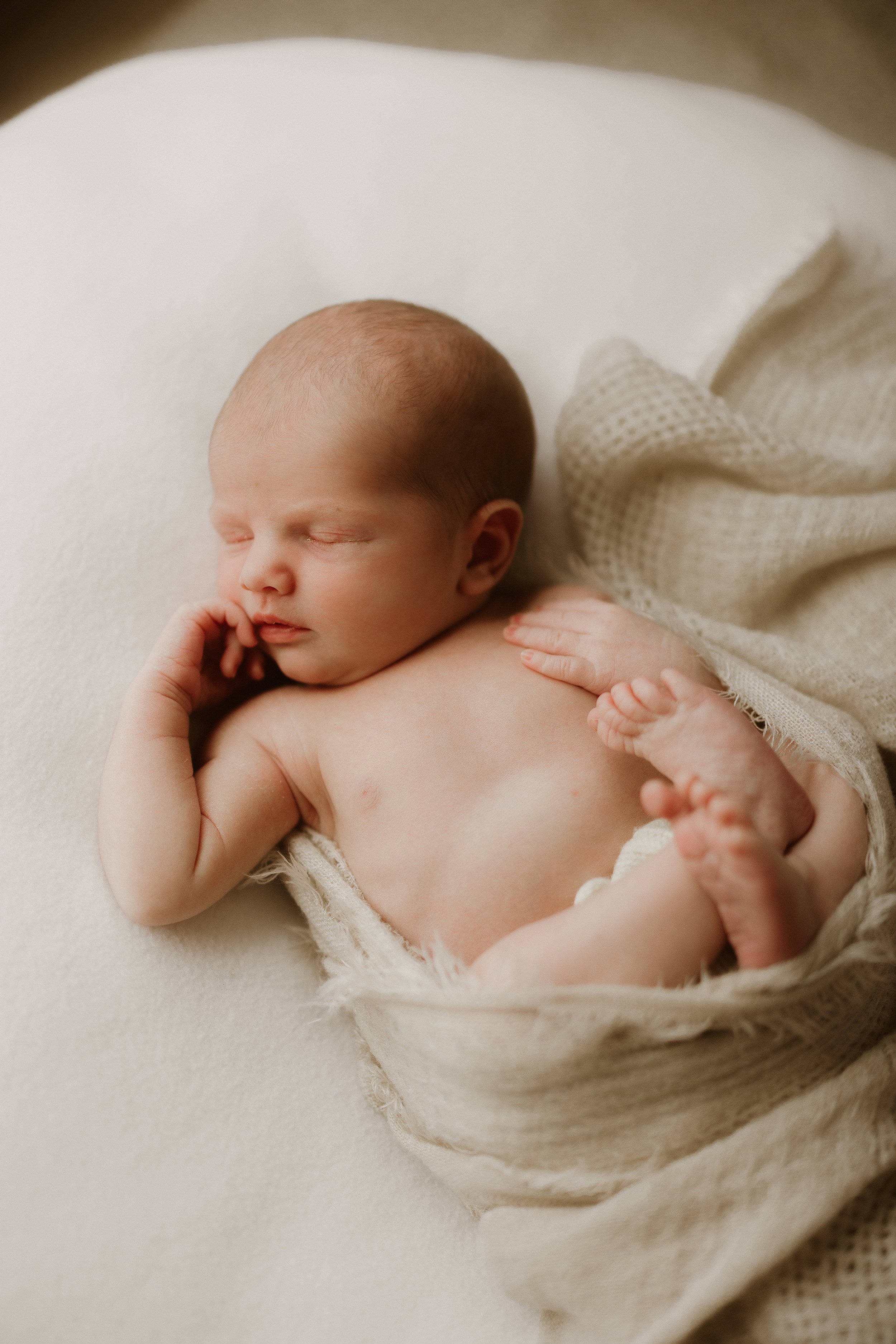 A newborn baby sleeping on a soft surface, wrapped in a beige knit blanket.