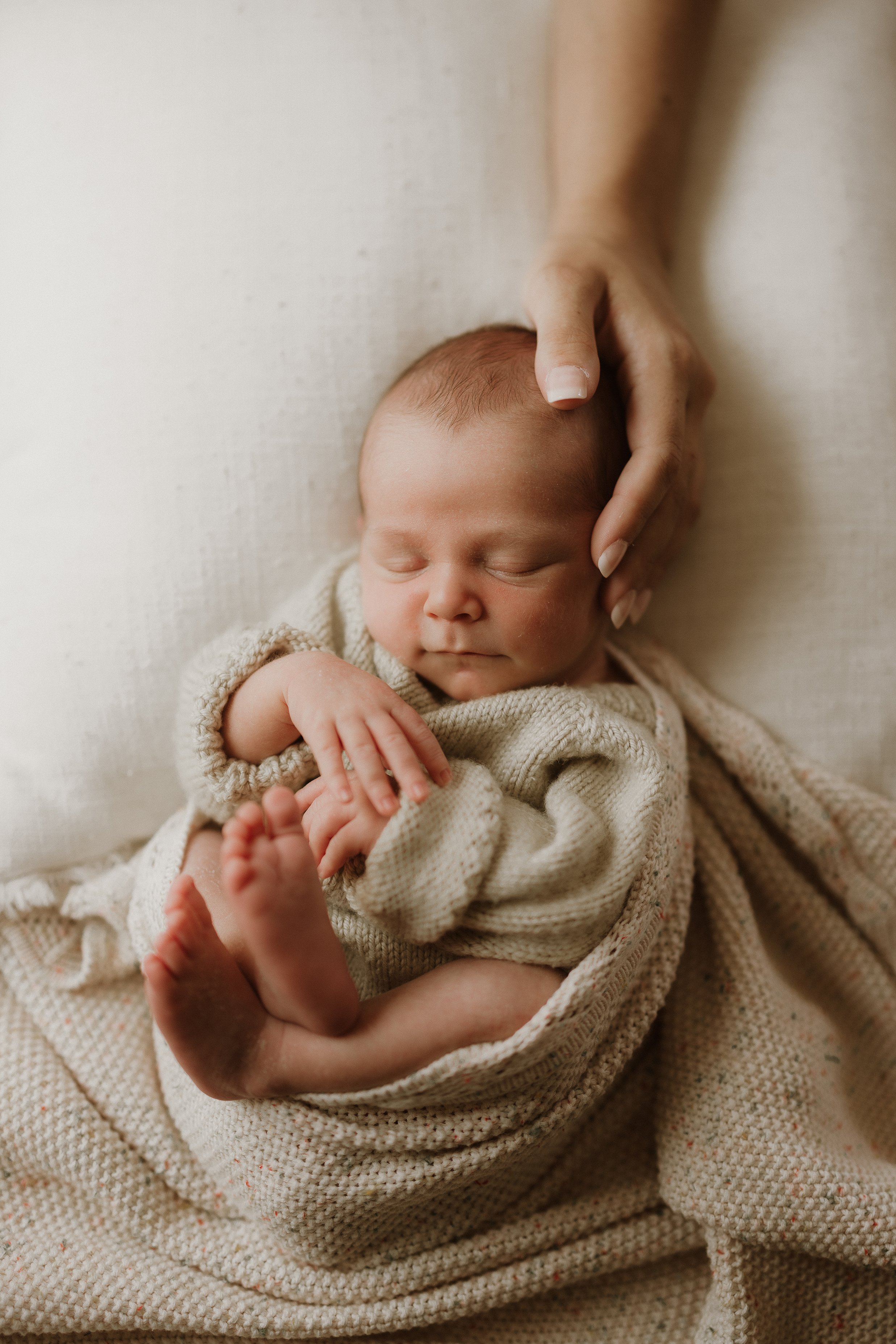 A sleeping newborn baby wrapped in a beige blanket with an adult hand gently touching the baby's head and shoulder.