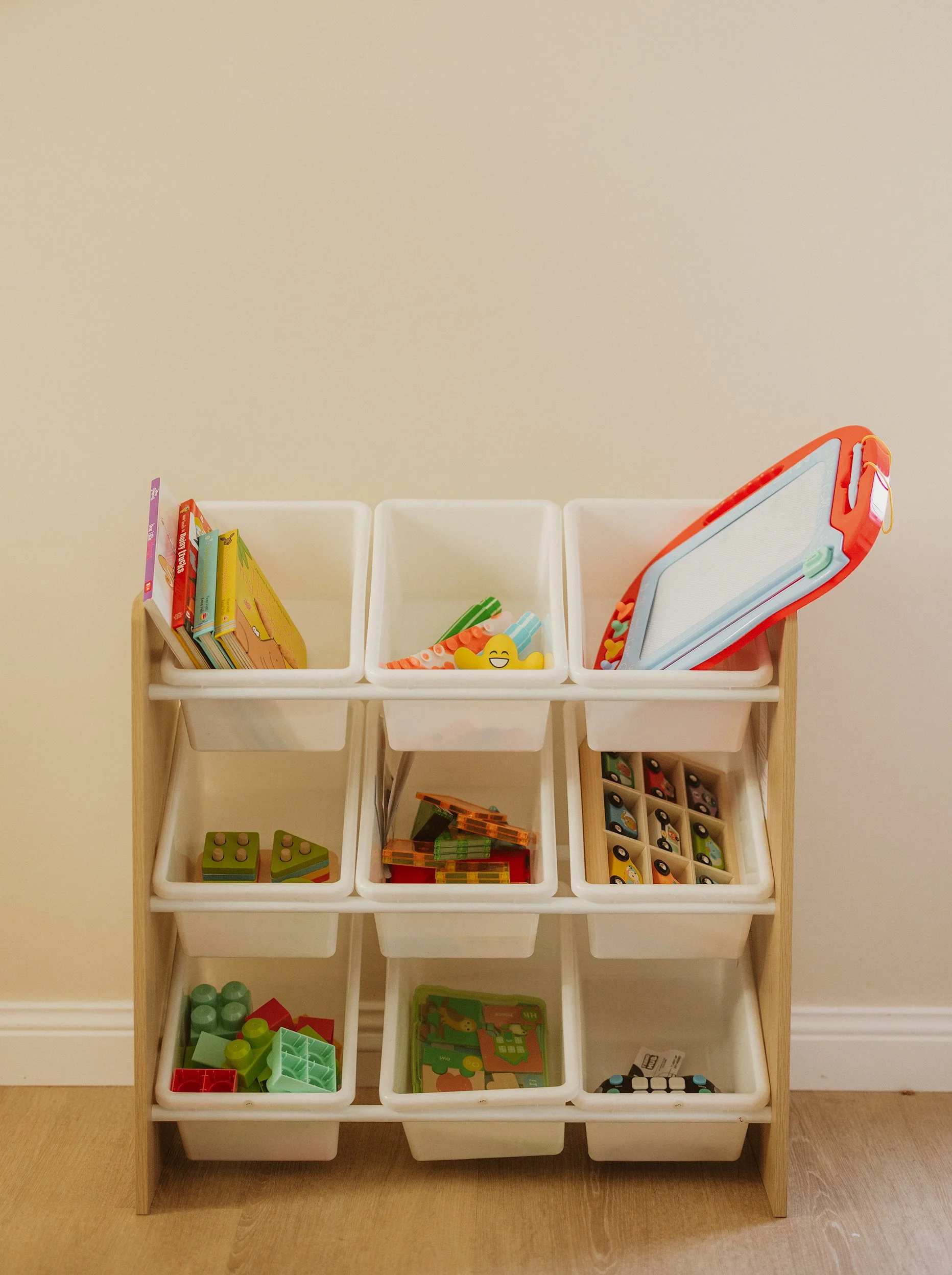Children's toy storage unit with nine white plastic bins filled with various toys, including books, building blocks, and toy cars, against a plain wall on a wooden floor.