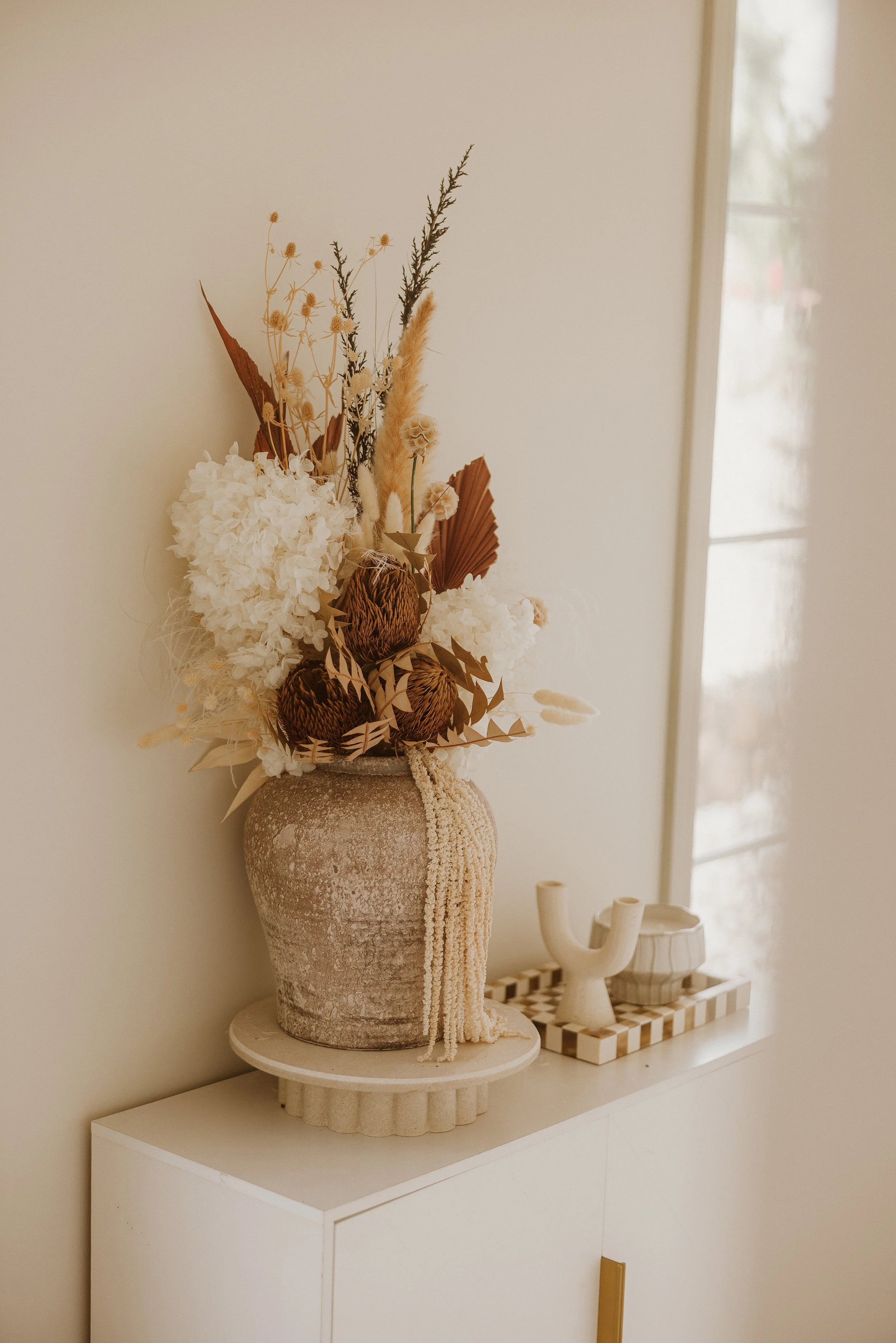 A decorative arrangement of dried flowers and plants in a textured ceramic vase on a white cabinet, with two small ceramic vases on a tray beside it, near a window.