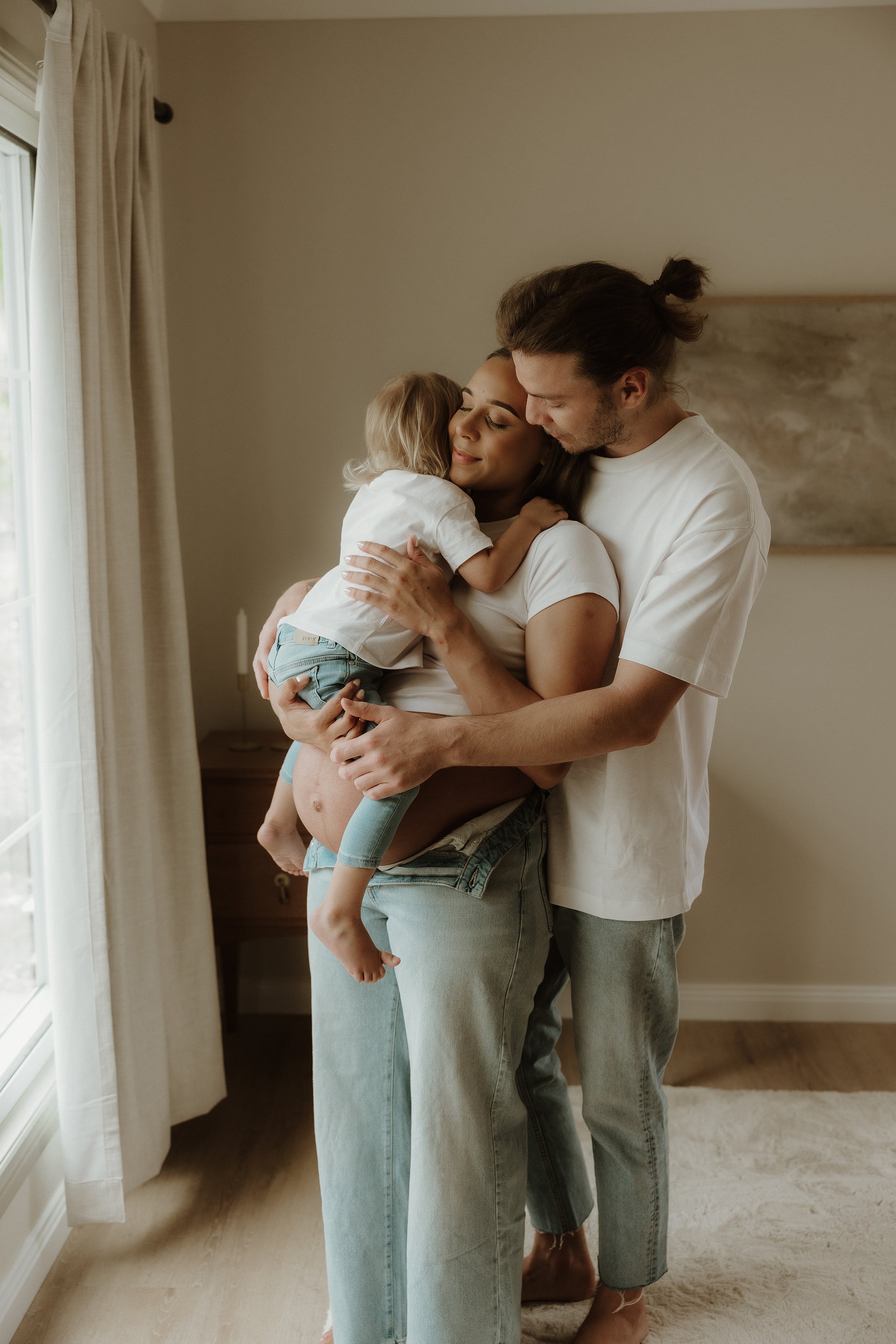 A pregnant woman hugging a young child while a man keeps close to them in a cozy living room.