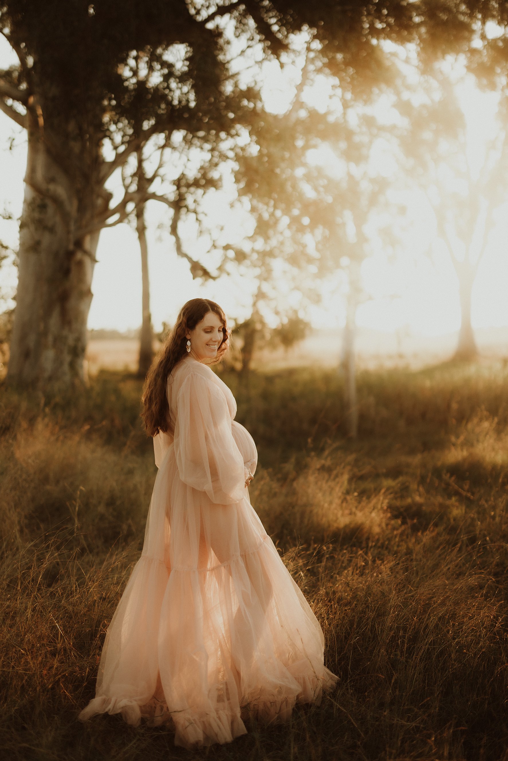A pregnant woman in a flowing, light-colored dress standing outdoors in a field at sunset, smiling and looking down at her belly.