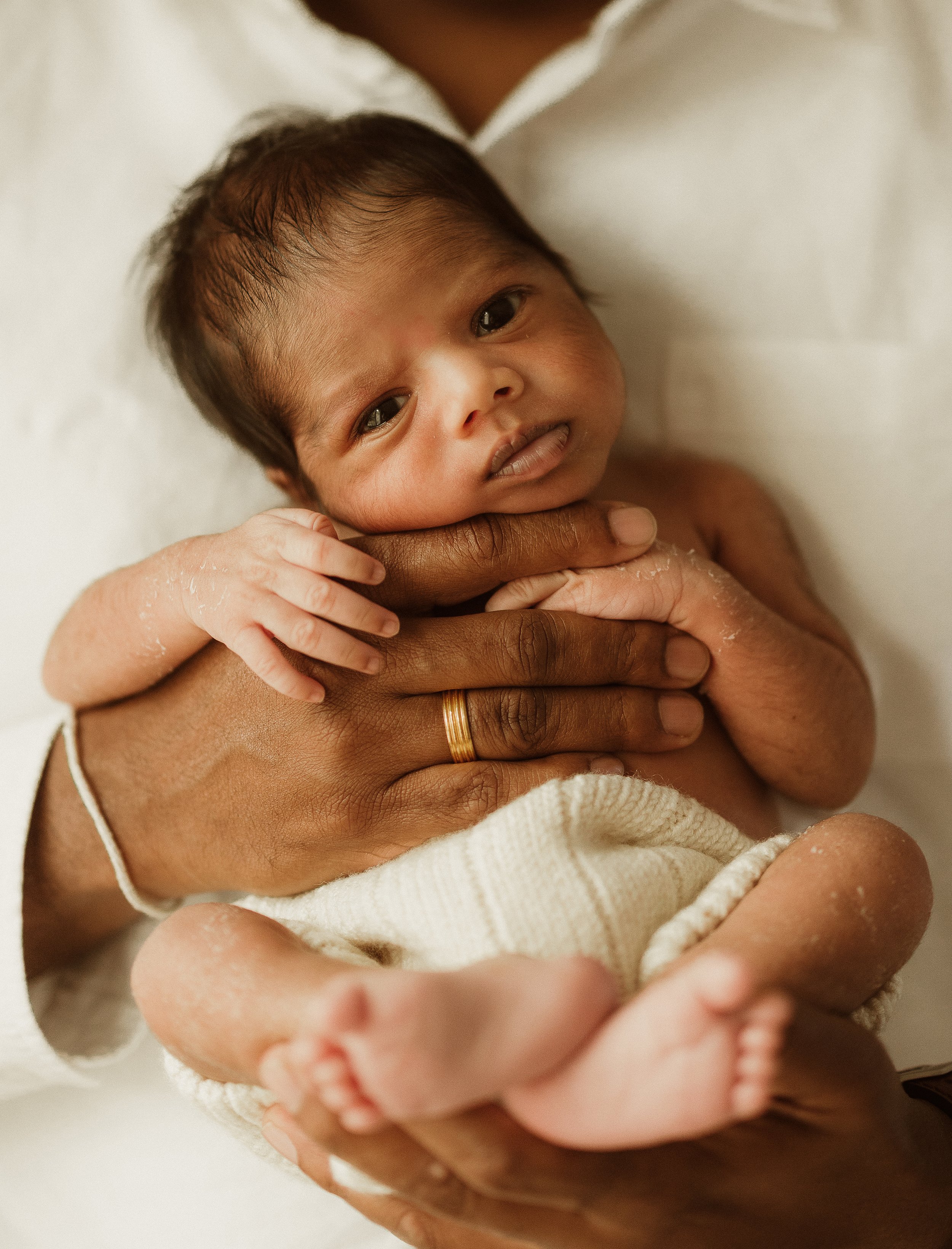 A newborn baby baby being gently held by an adult with a gold ring on their finger. The baby has dark hair and is lying on a beige blanket, looking at the camera with a curious expression.