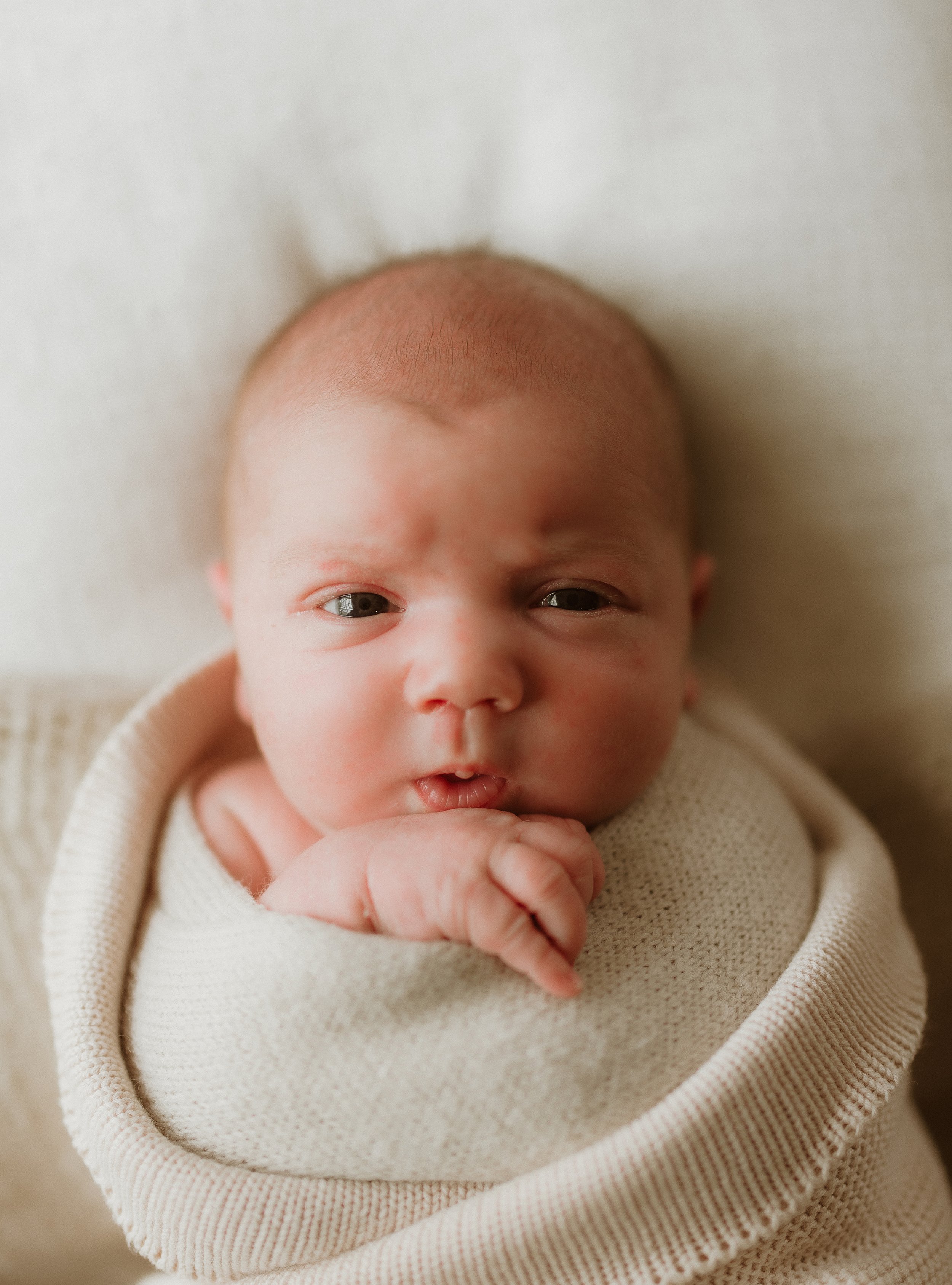 Close-up of a newborn baby with light skin and gray eyes, wrapped in a beige blanket, resting on a soft surface, looking at the camera.