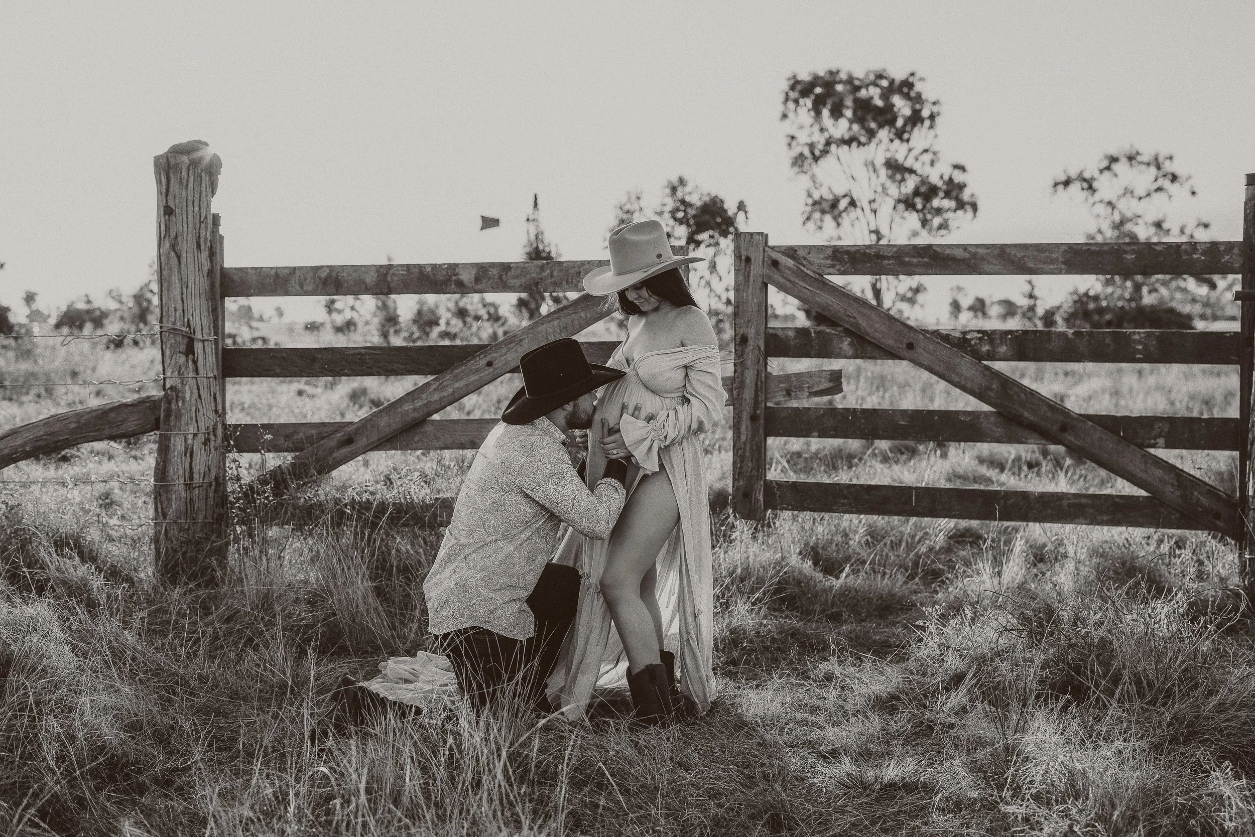 A woman in a flowing dress and cowboy boots, pregnant, is posing outdoors by a wooden fence while a man kneels and kisses her belly. Both are wearing wide-brimmed hats, and the scene is in black and white with a rural background.