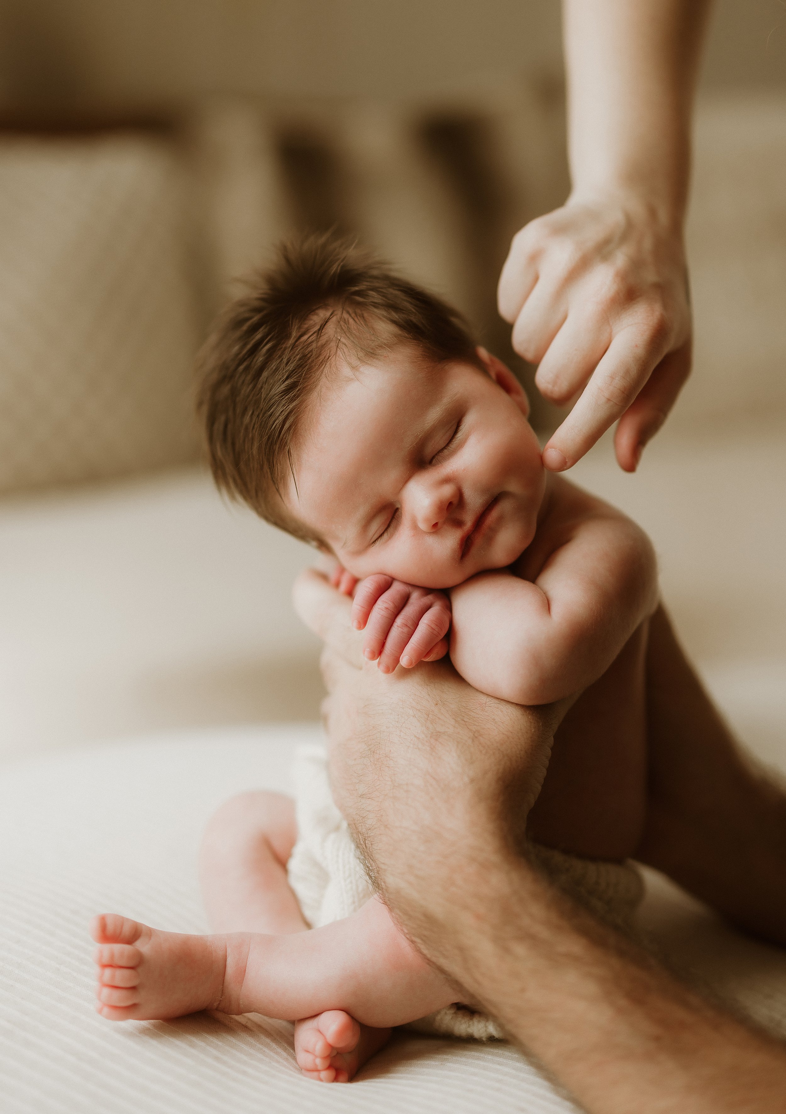 A close-up of a baby with closed eyes, resting on an adult's hand, with another hand gently touching the baby's nose.