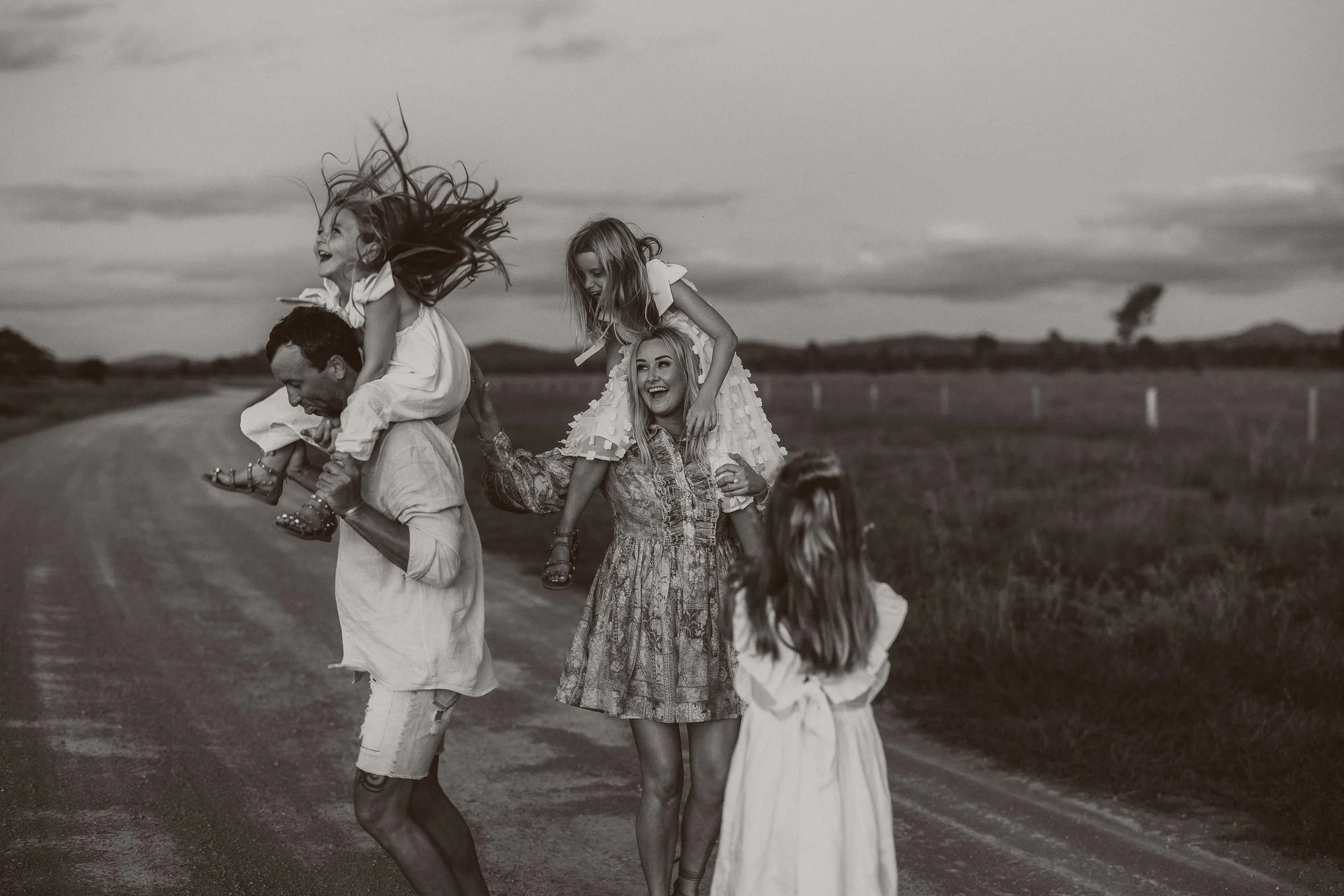 A group of children and a woman are outdoors on a dirt road, playing and having fun, with some girls sitting on the woman's shoulders, in a joyful moment.
