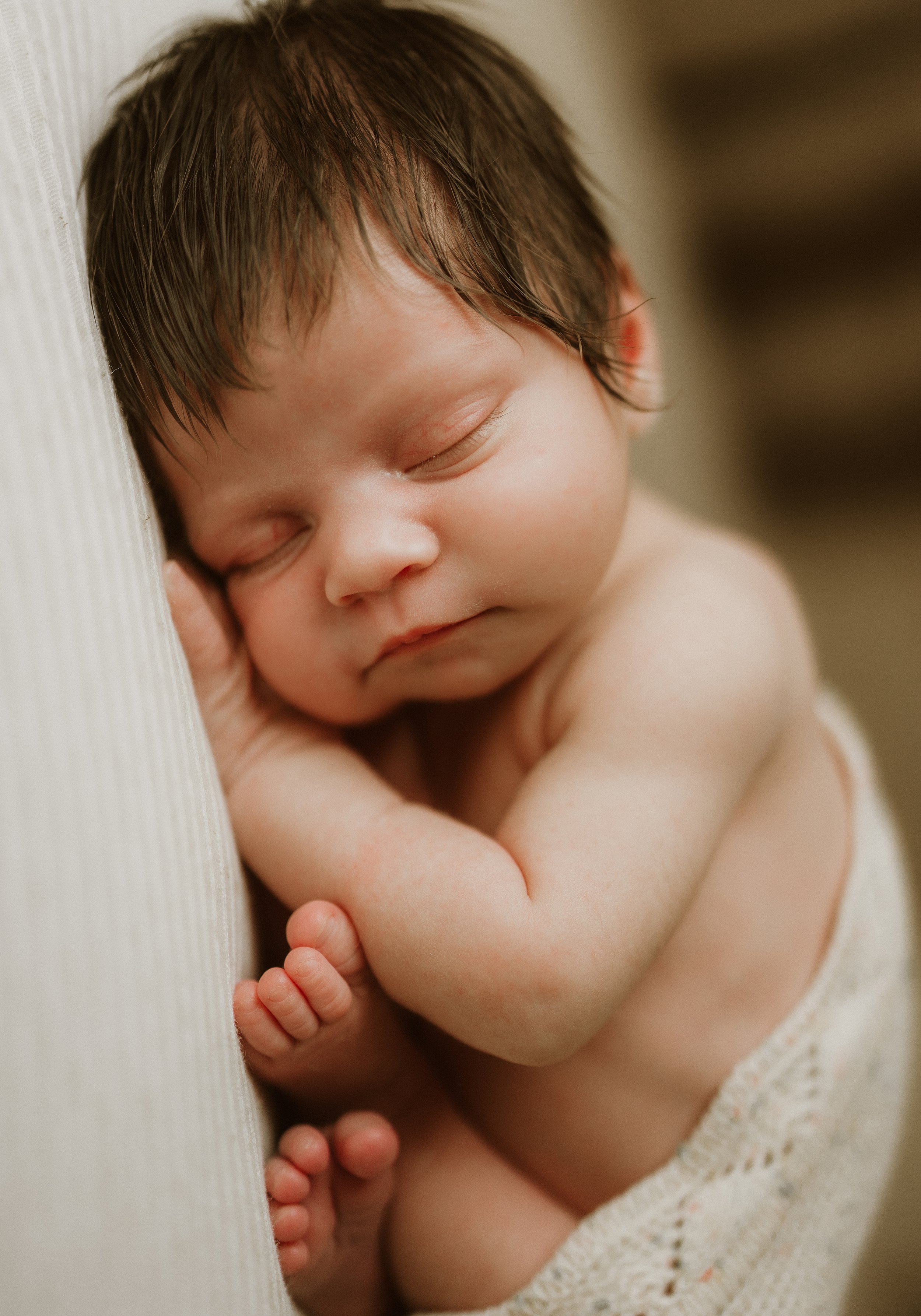 A sleeping newborn baby with brown hair resting peacefully on a soft surface, curled up with arms and legs close to the body, wearing a light knitted cloth.