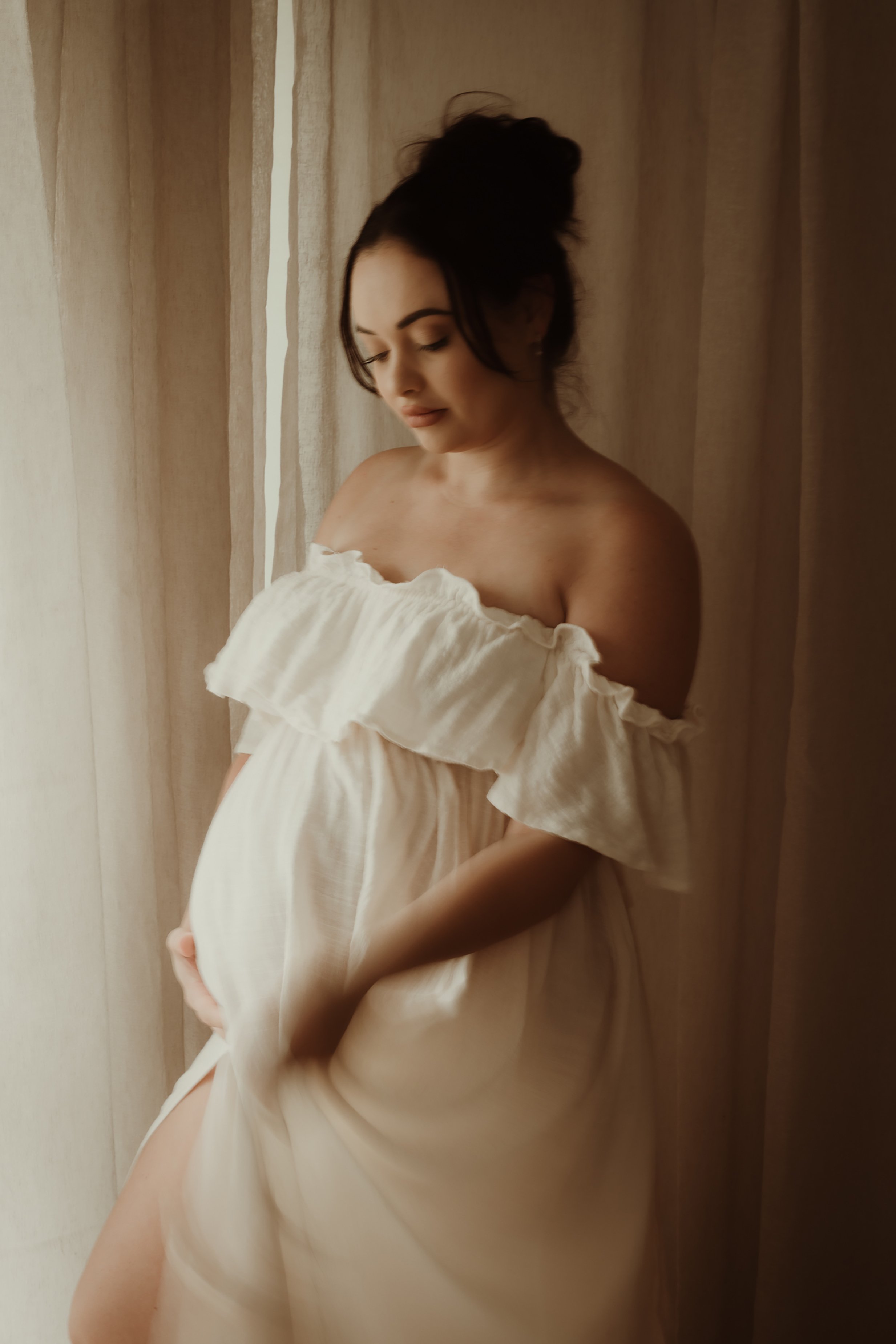 A pregnant woman in an off-the-shoulder cream-colored dress standing near a window with curtains, gently cradling her baby bump and looking down.