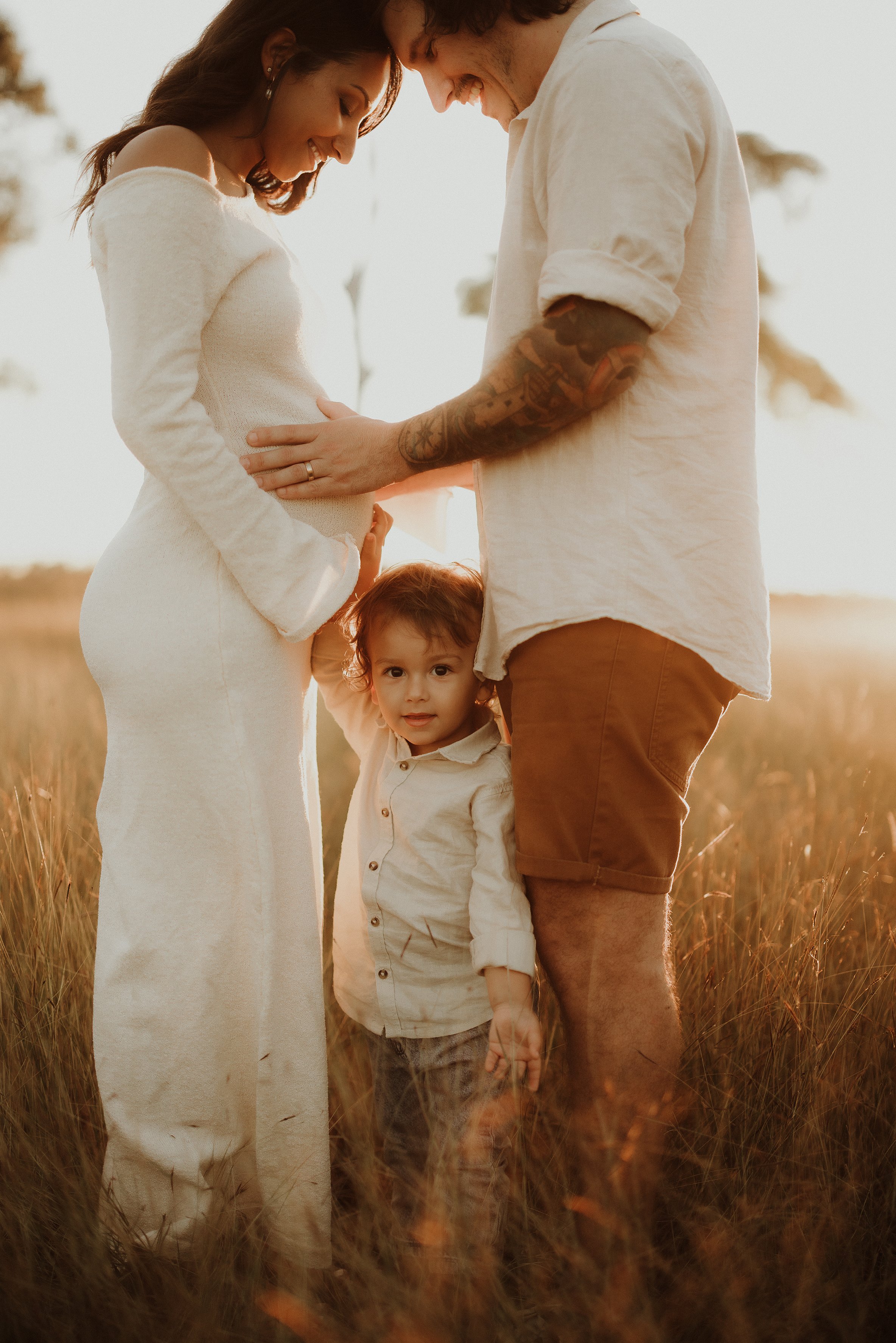 A pregnant woman and a man standing close together holding her belly, with a young boy standing between them touching her belly, in a grassy field during sunset.