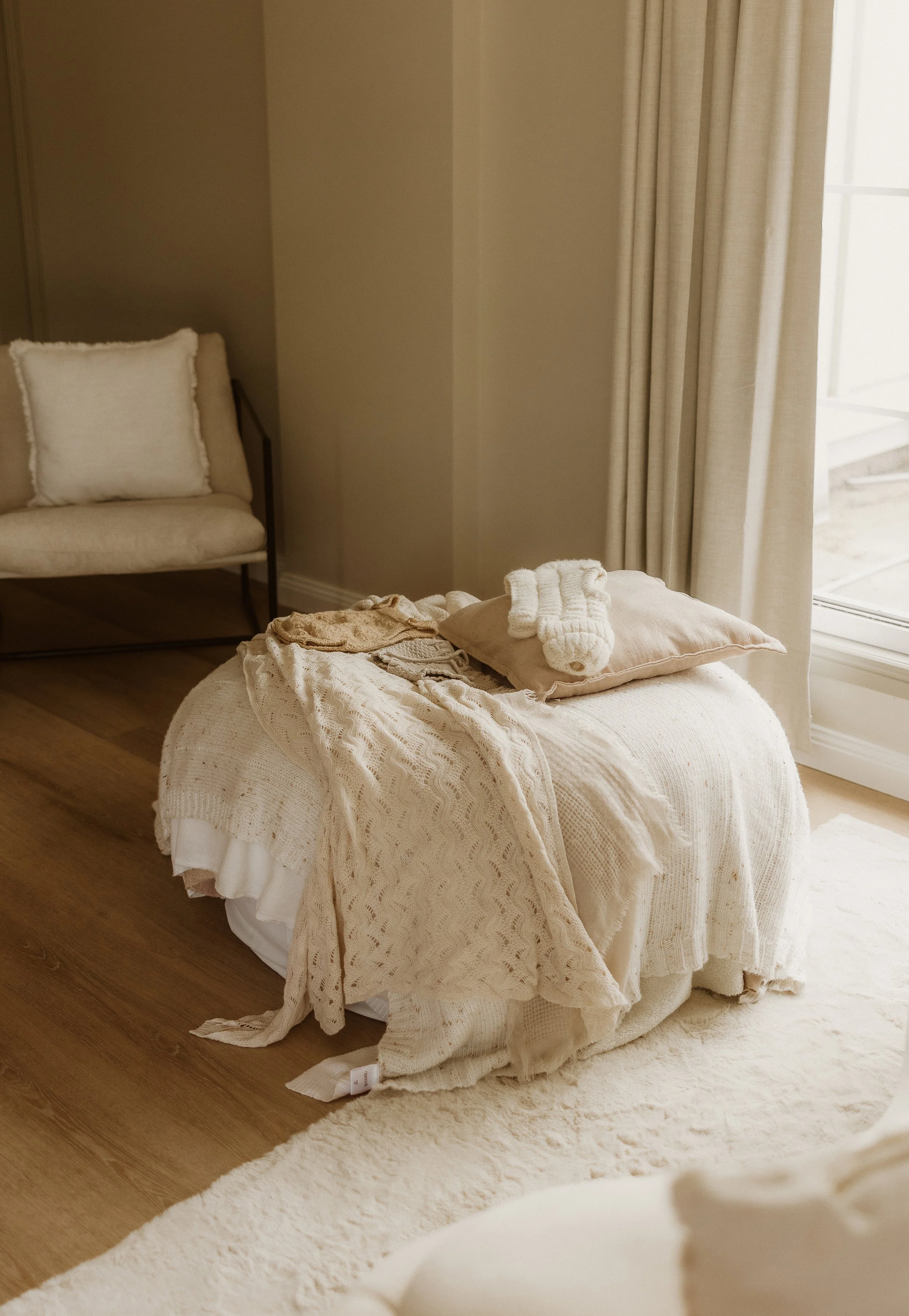 A cozy bedroom corner with a white textured bedspread, beige pillows, several pieces of clothing, and knit accessories on a bed near a window with cream curtains.