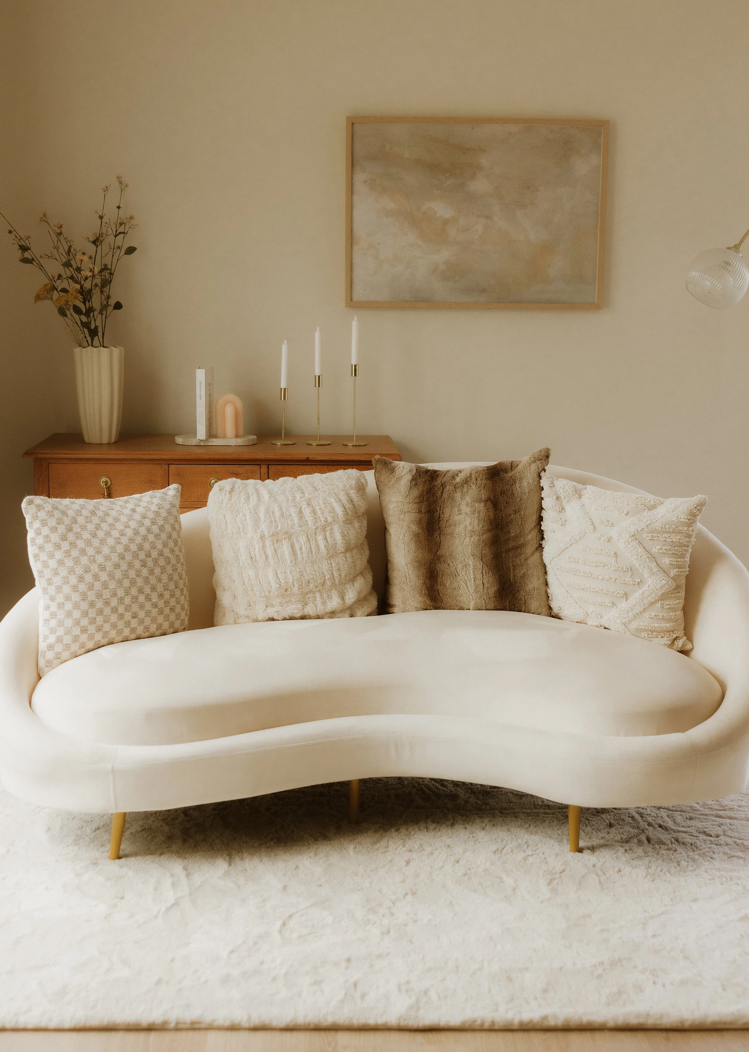 Living room with a white curved sofa adorned with four textured throw pillows, a beige plush rug, and a wooden sideboard with decorative candles, a vase with dried flowers, and abstract art on the wall.