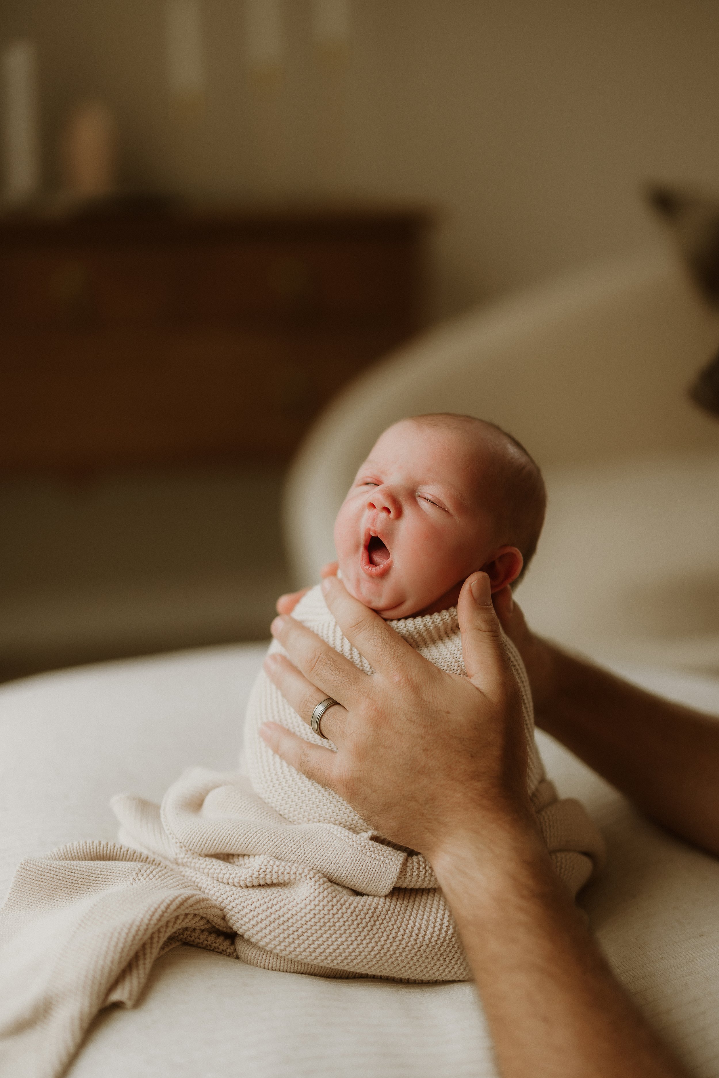 A newborn baby with eyes closed and mouth open, being held gently by an adult's hands, lying on a soft surface.